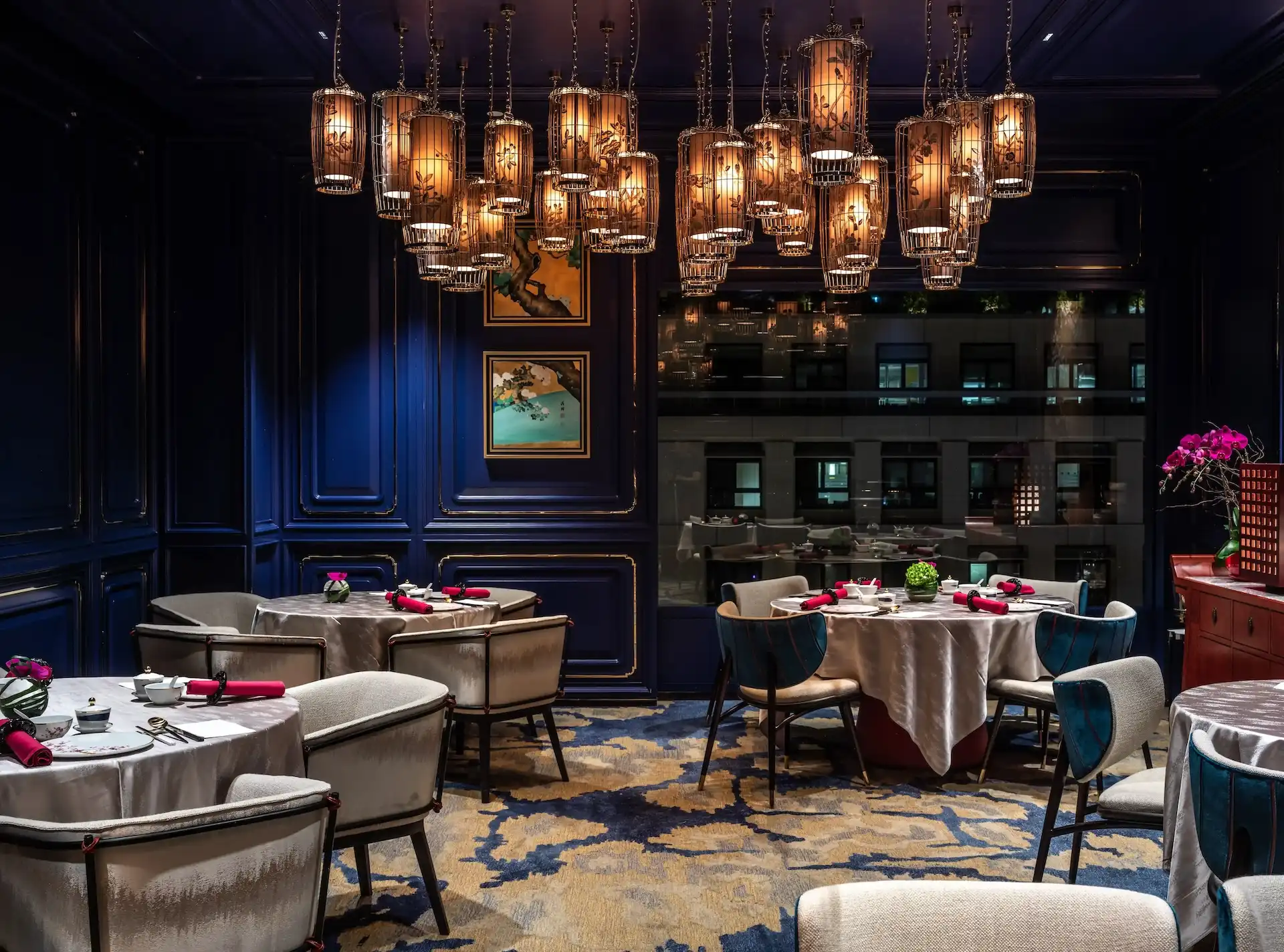 Dining room at Man Wah Mandarin Oriental Hong Kong featuring clustered lantern chandeliers with warm illumination, navy blue panelled walls with gold trim, round dining tables with white tablecloths and red napkins, patterned carpet in blue and gold, and framed artwork.