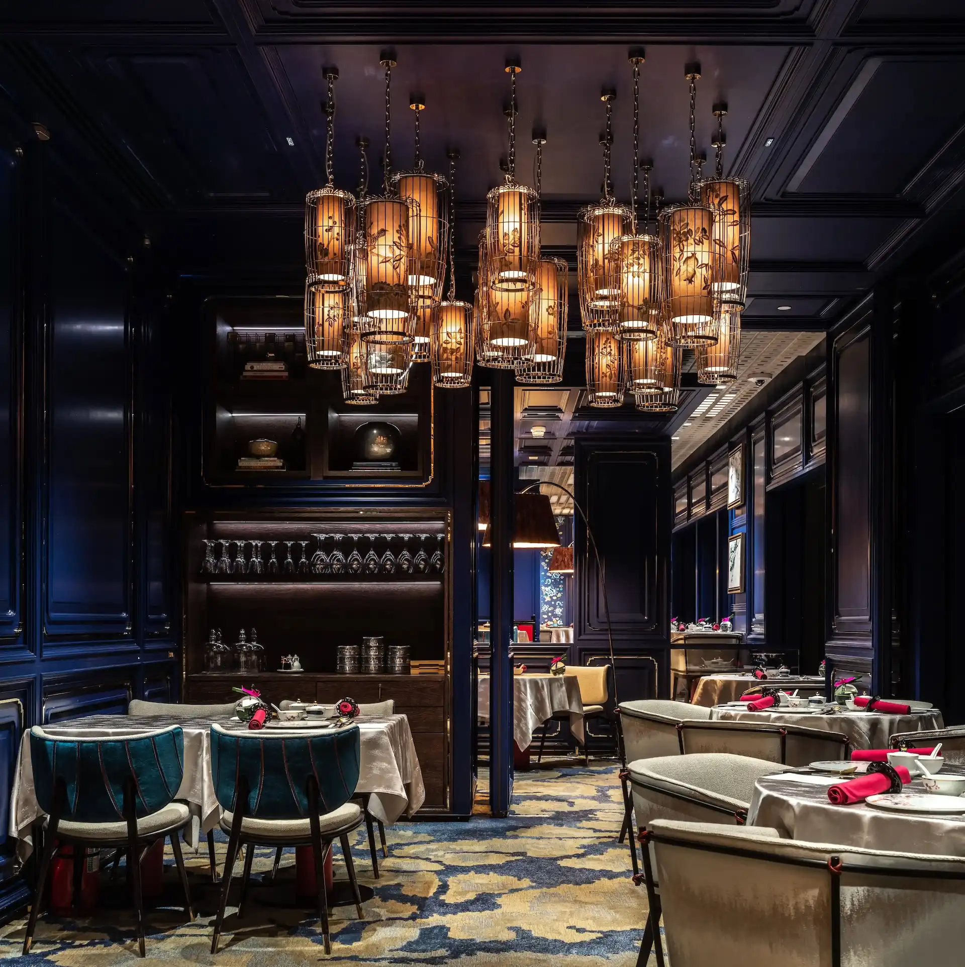 Interior of Man Wah Mandarin Oriental Hong Kong featuring lantern chandeliers with warm illumination, navy panelled walls with gold trim, open shelving with glassware, round dining tables dressed in white tablecloths and red napkins, and blue and gold patterned carpet.