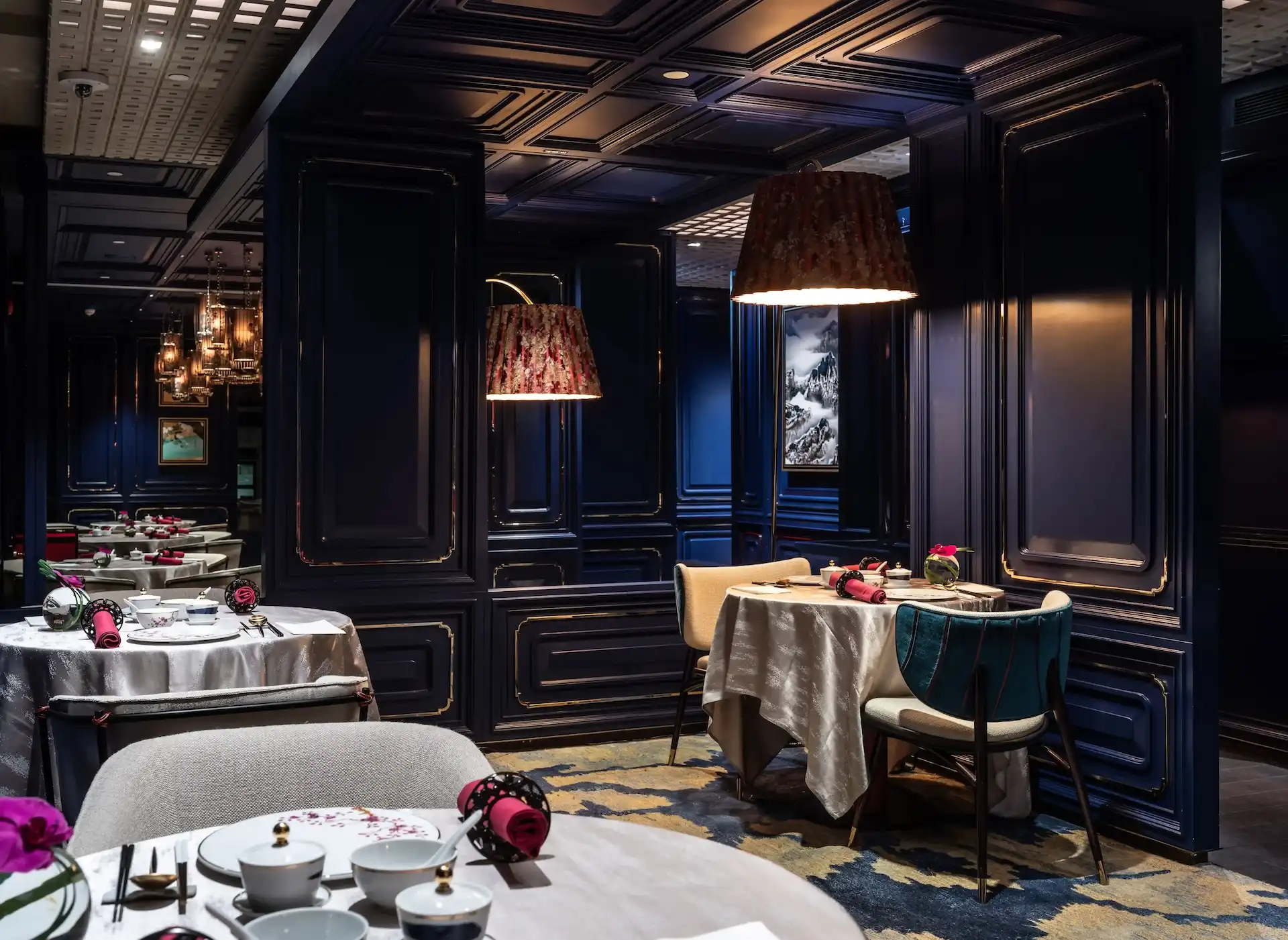 Intimate dining area at Man Wah Mandarin Oriental Hong Kong featuring navy panelled walls with gold detailing, silk lampshades with warm glow, round tables dressed in white tablecloths, upholstered chairs in neutral and teal tones, and patterned blue and gold carpet.