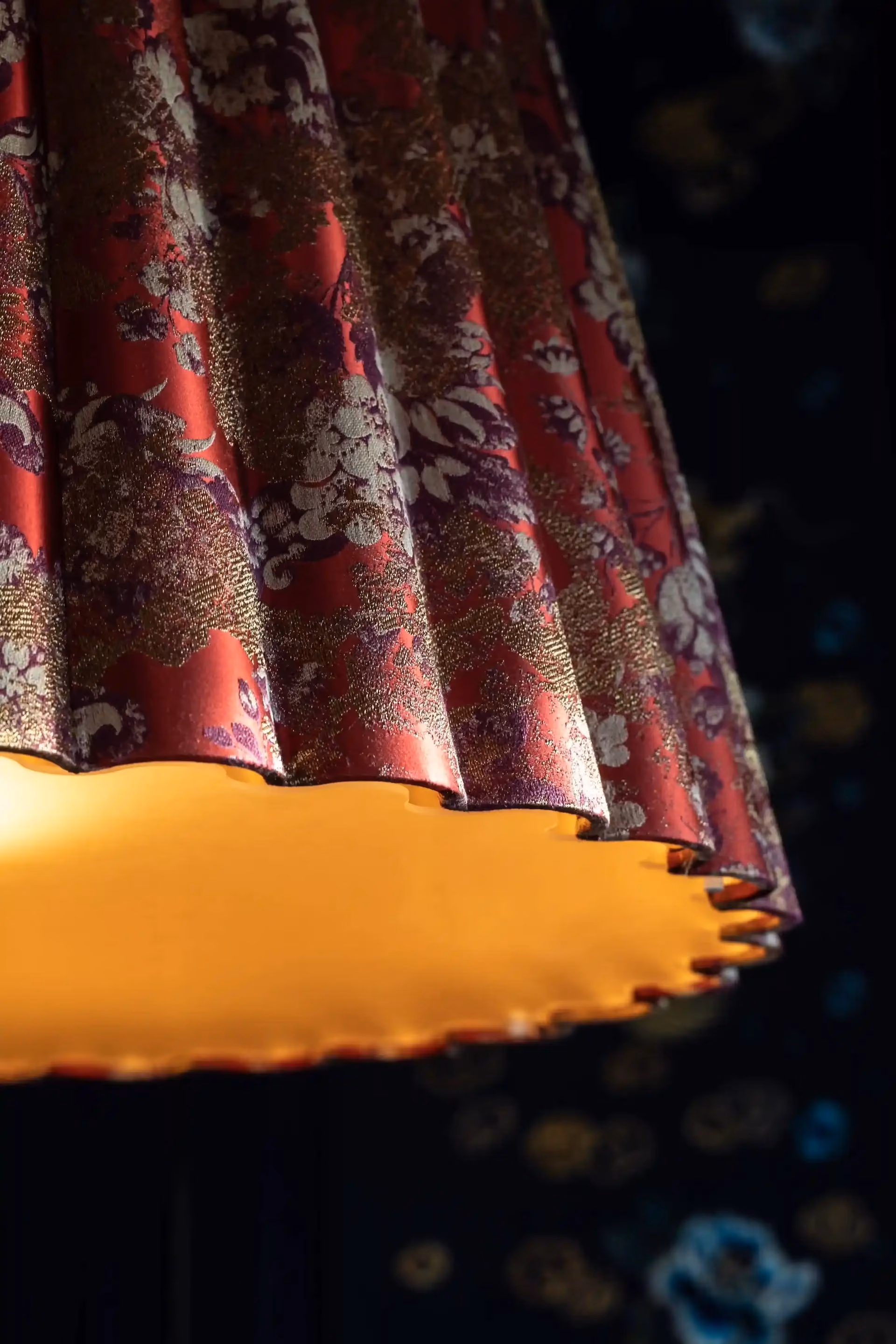 Close up detail of silk lampshade at Man Wah Mandarin Oriental Hong Kong featuring red and gold floral fabric with scalloped edge and warm internal illumination against dark patterned backdrop.