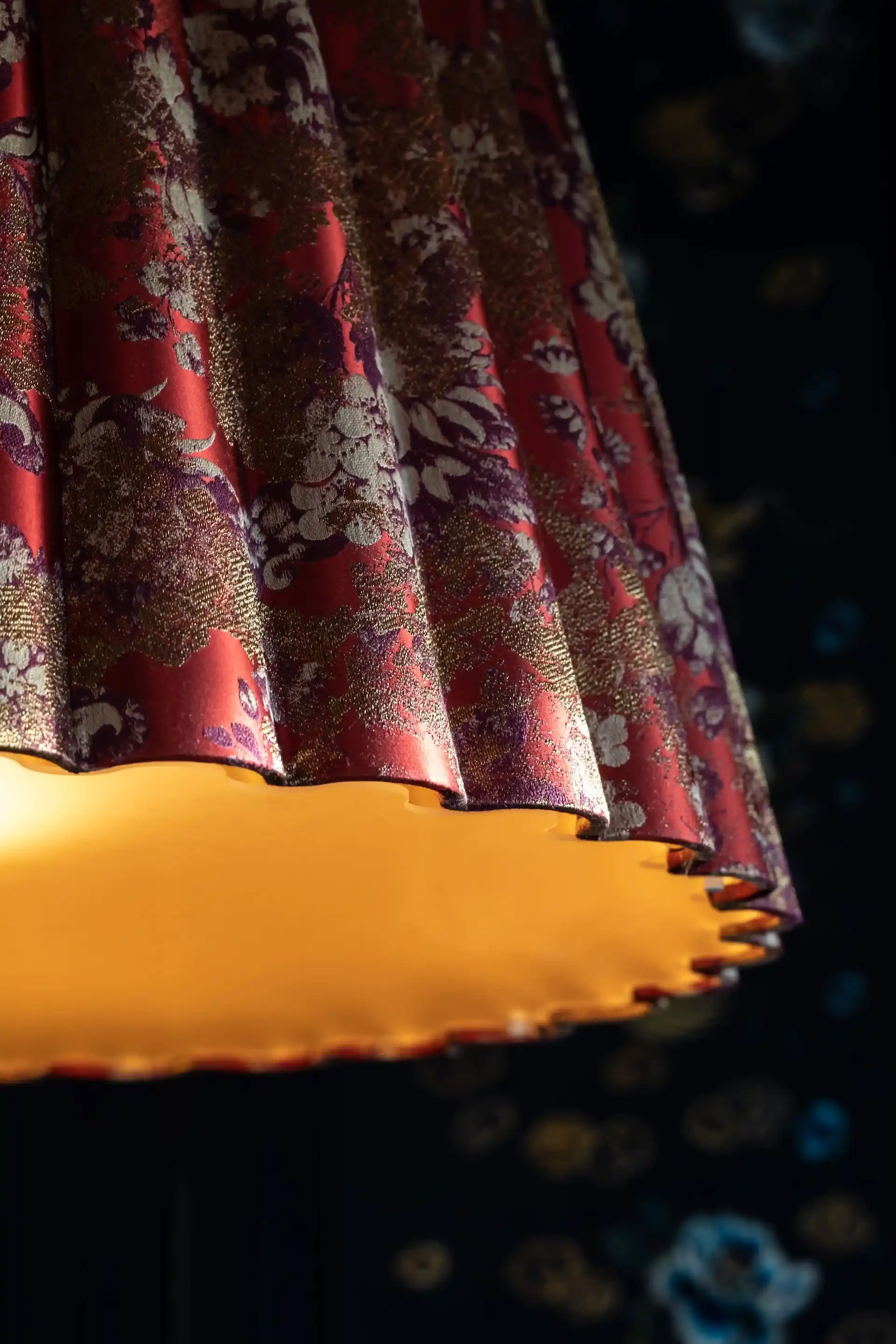 Close up detail of silk lampshade at Man Wah Mandarin Oriental Hong Kong featuring red and gold floral fabric with scalloped edge and warm internal illumination against dark patterned backdrop.