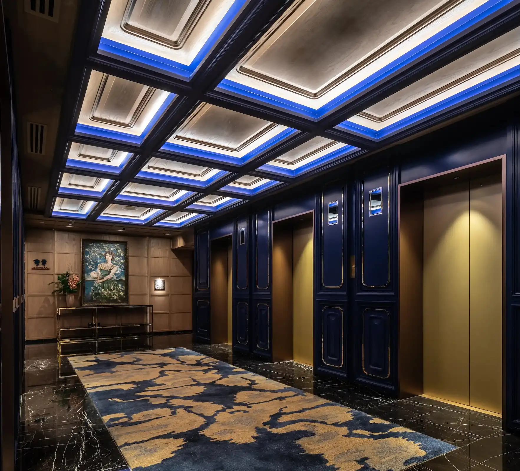Lift lobby at Man Wah Mandarin Oriental Hong Kong featuring deep navy panelled walls with gold trim, coffered ceiling with blue cove lighting, black marble flooring, patterned carpet runner in blue and gold, and framed artwork with console table at corridor end.