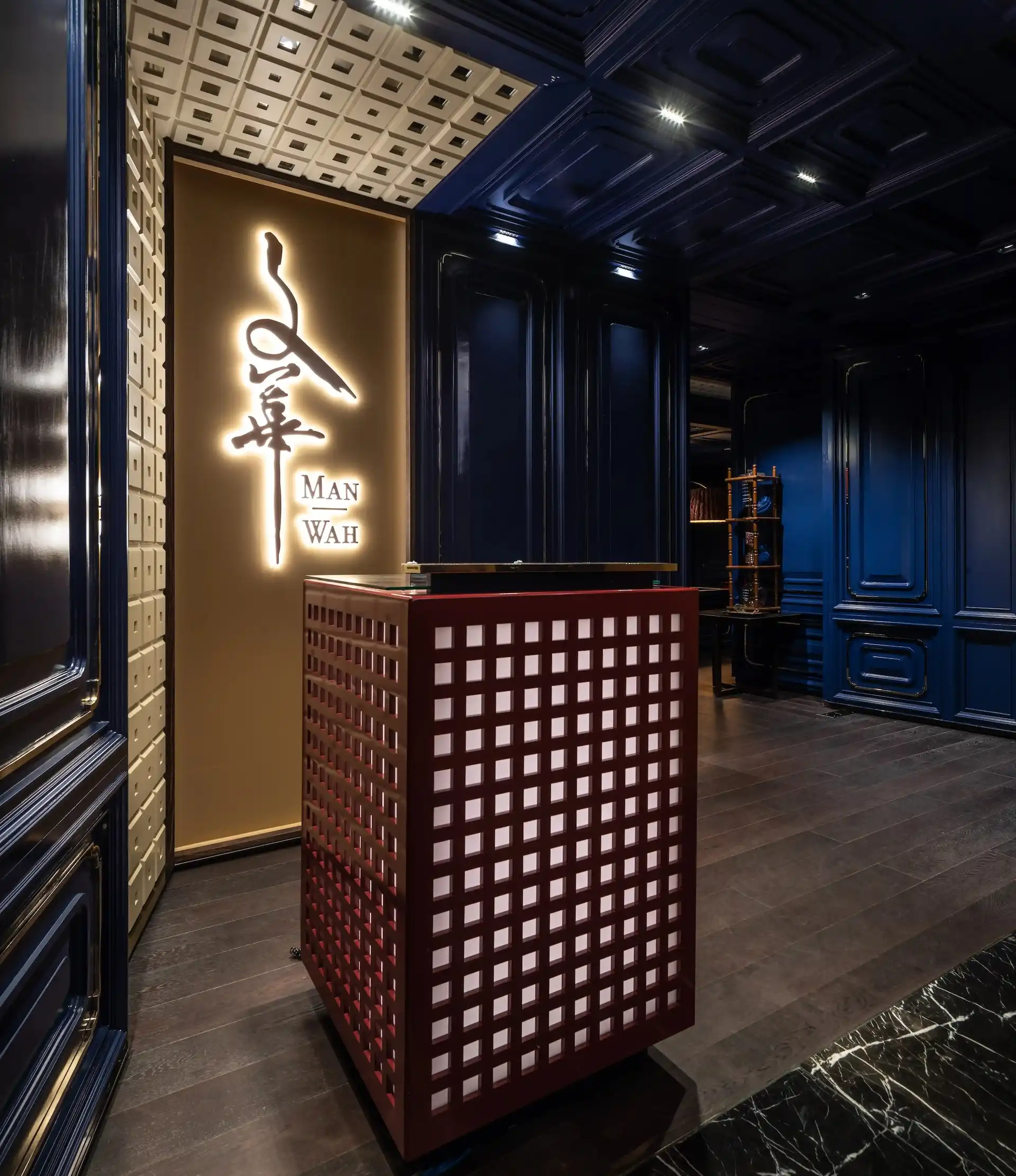 Entrance at Man Wah Mandarin Oriental Hong Kong featuring illuminated Chinese calligraphy logo on bronze wall, red lattice host podium with glass top, navy blue panelled walls with gold detailing, coffered ceiling, and dark timber flooring.