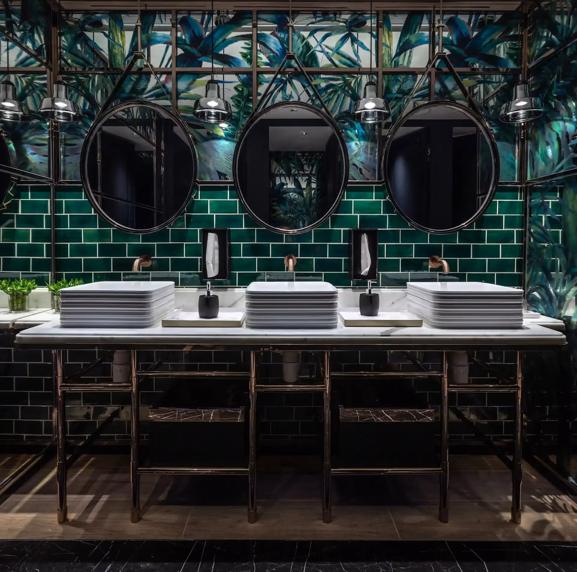 Washroom vanity at Man Wah Mandarin Oriental Hong Kong featuring green subway tile walls, botanical mural panels, triple round mirrors with industrial light fittings, stacked rectangular basins on marble countertop, and dark metal framed console base.