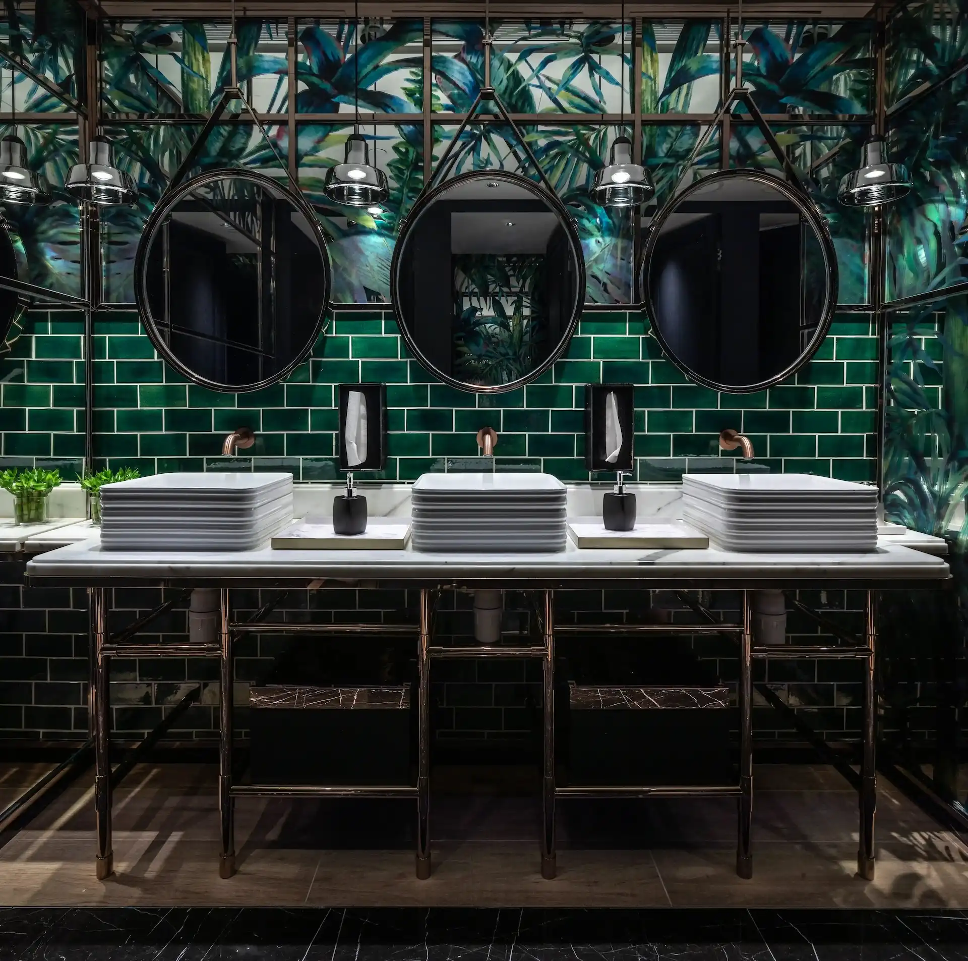 Washroom vanity at Man Wah Mandarin Oriental Hong Kong featuring green subway tile walls, botanical mural panels, triple round mirrors with industrial light fittings, stacked rectangular basins on marble countertop, and dark metal framed console base.