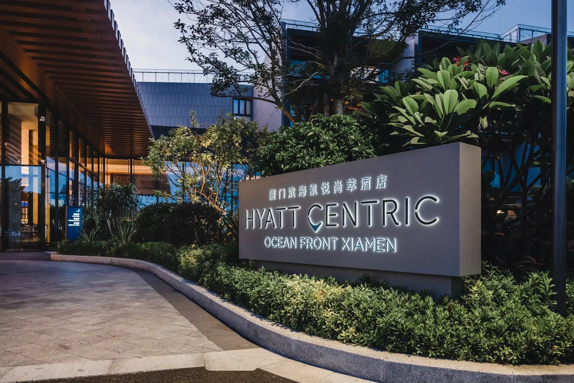 Entrance signage at Hyatt Centric Oceanfront Xiamen China featuring illuminated hotel logo and lettering set within landscaped greenery, modern low rise façade, and covered drop off area at dusk.