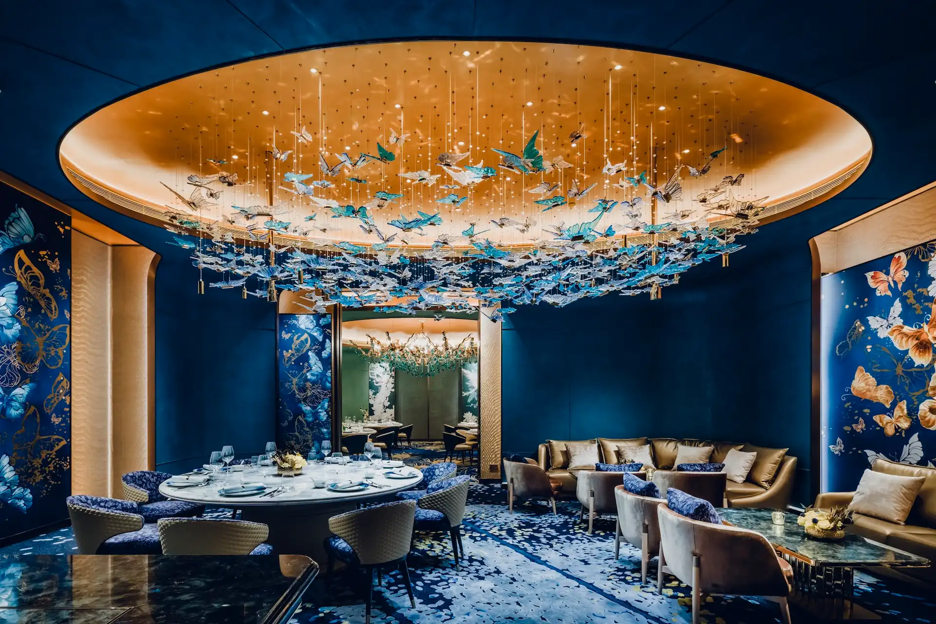 Grand Imperial Court Tapestry Butterfly Wings private dining suite at MGM Macao featuring illuminated oval ceiling with suspended butterfly chandelier deep blue curved wall panels bespoke butterfly artwork round dining table and adjoining lounge seating within a luxury Chinese restaurant interior design