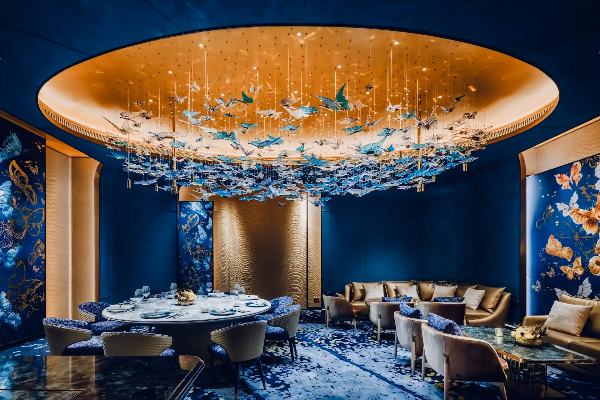 Grand Imperial Court Tapestry Butterfly Wings private dining interior at MGM Macao featuring illuminated oval ceiling recess with suspended butterfly chandelier deep blue wall panels bespoke butterfly artwork round dining table lounge seating and patterned carpet within a luxury Chinese restaurant design