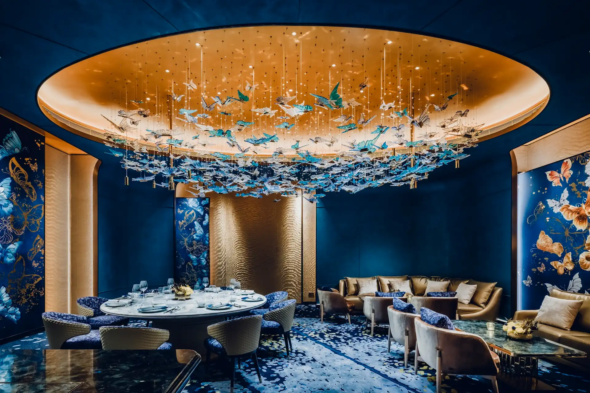 Grand Imperial Court Tapestry Butterfly Wings private dining interior at MGM Macao featuring illuminated oval ceiling recess with suspended butterfly chandelier deep blue wall panels bespoke butterfly artwork round dining table lounge seating and patterned carpet within a luxury Chinese restaurant design