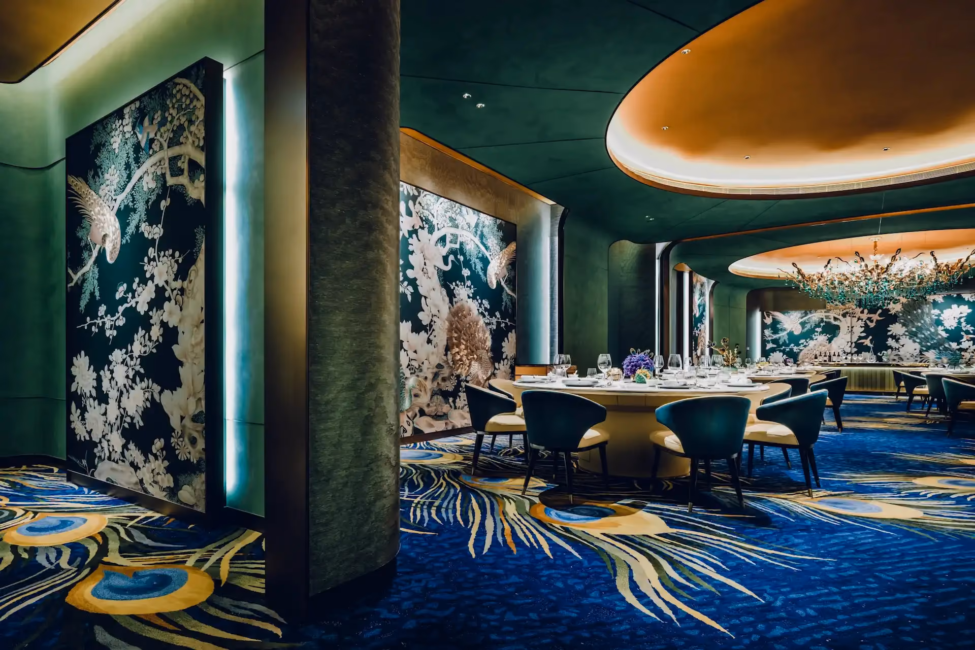 Grand Imperial Court Tapestry Of Feathers private dining room at MGM Macao featuring round banquet table teal upholstered chairs large scale bird and floral wall murals illuminated ceiling recess crystal chandelier and bespoke peacock feather patterned carpet within a luxury Chinese restaurant interior design