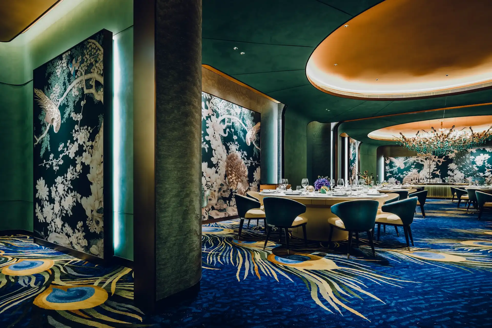 Grand Imperial Court Tapestry Of Feathers private dining room at MGM Macao featuring round banquet table teal upholstered chairs large scale bird and floral wall murals illuminated ceiling recess crystal chandelier and bespoke peacock feather patterned carpet within a luxury Chinese restaurant interior design