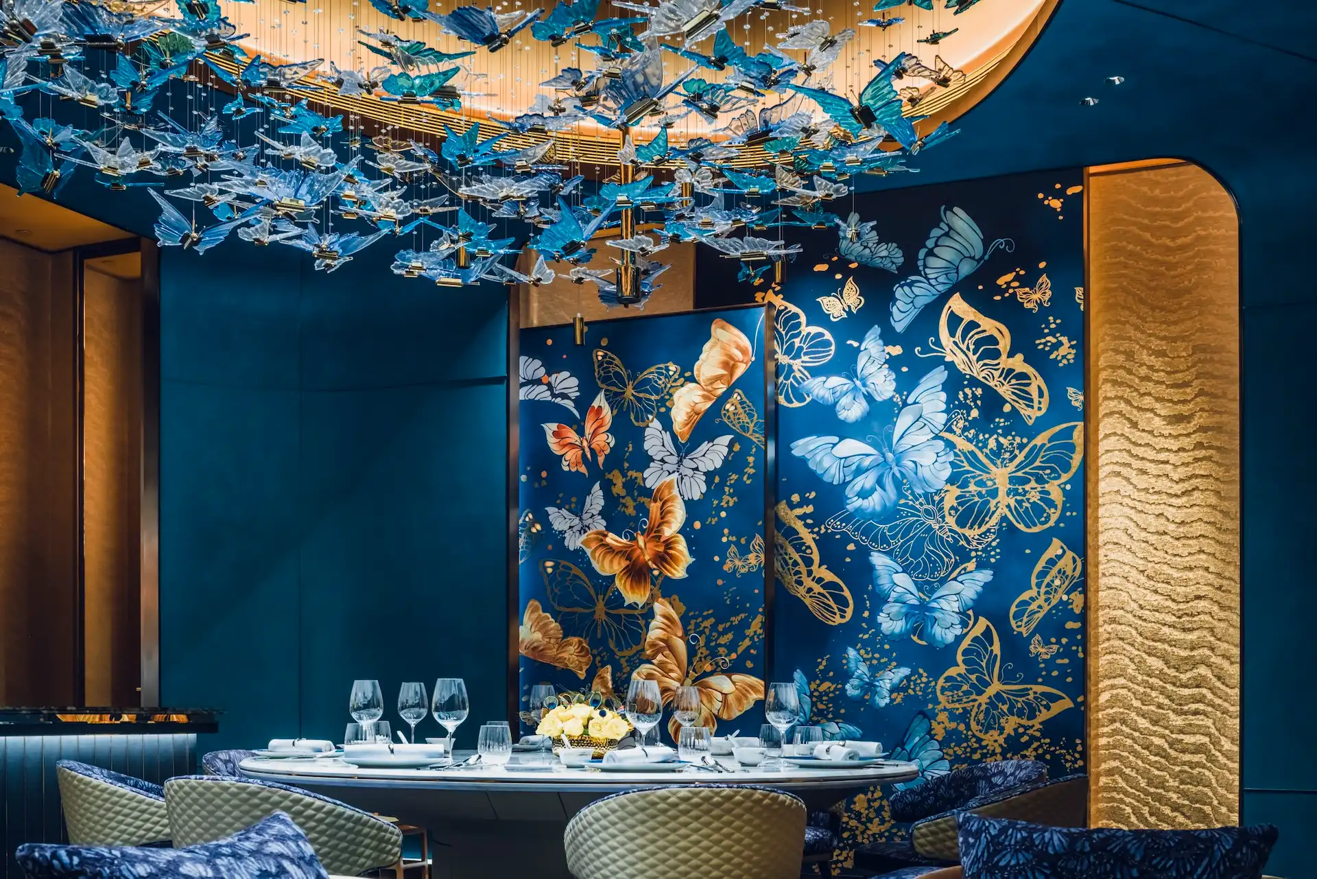 Grand Imperial Court private dining detail at MGM Macao featuring bespoke butterfly wall panels in blue and gold tones circular dining table crystal glassware and suspended butterfly chandelier within a luxury Chinese restaurant interior design