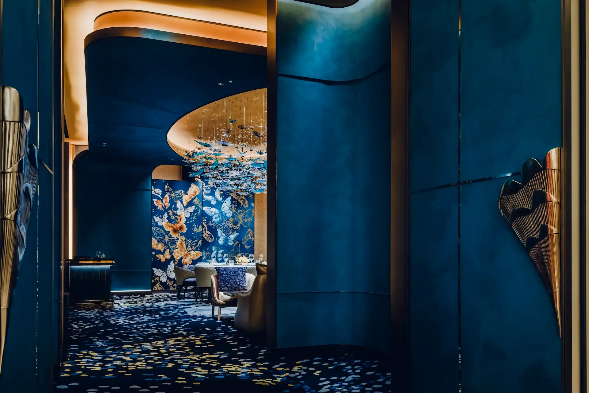 Grand Imperial Court Tapestry Butterfly Wings private dining interior at MGM Macao featuring deep blue wall panels bespoke butterfly artwork sculptural brass door handles custom patterned carpet and suspended butterfly light installation within a luxury Chinese restaurant design
