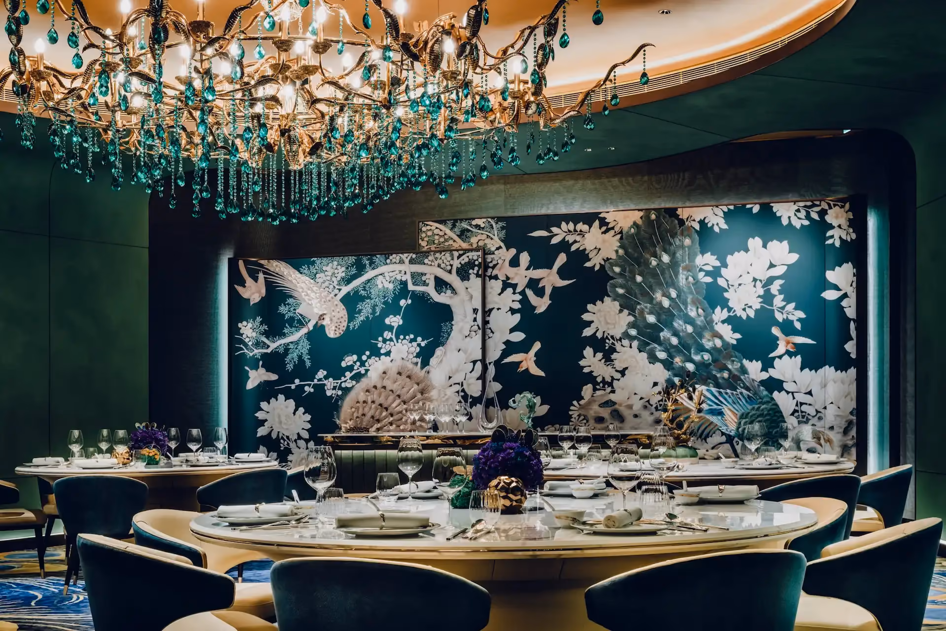 Grand Imperial Court Tapestry Of Feathers chandelier detail at MGM Macao featuring sculptural crystal and emerald glass lighting suspended beneath illuminated ceiling recess with round banquet tables bespoke bird and floral wall murals and teal upholstered dining chairs within a luxury Chinese restaurant interior design