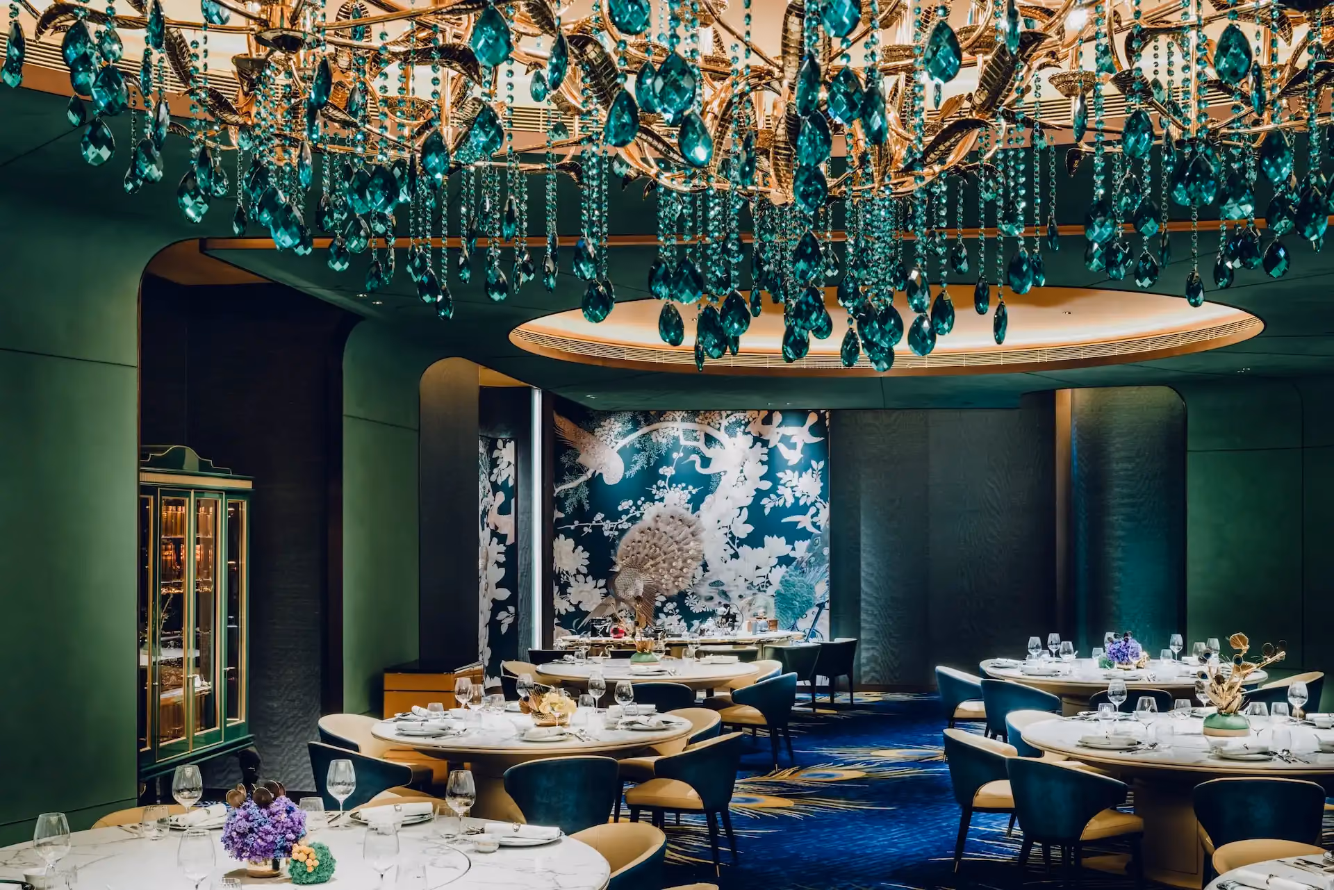 Grand Imperial Court Tapestry Of Feathers dining interior at MGM Macao featuring emerald crystal chandelier beneath illuminated circular ceiling recess round banquet tables teal upholstered chairs bespoke bird and floral wall murals and peacock feather patterned carpet within a luxury Chinese restaurant design