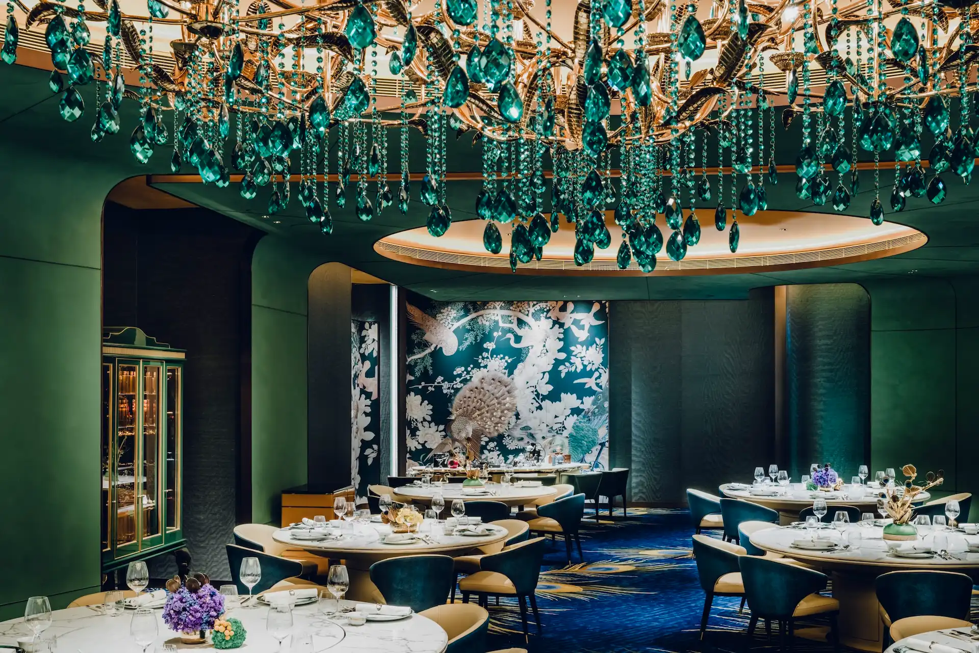 Grand Imperial Court Tapestry Of Feathers dining interior at MGM Macao featuring emerald crystal chandelier beneath illuminated circular ceiling recess round banquet tables teal upholstered chairs bespoke bird and floral wall murals and peacock feather patterned carpet within a luxury Chinese restaurant design