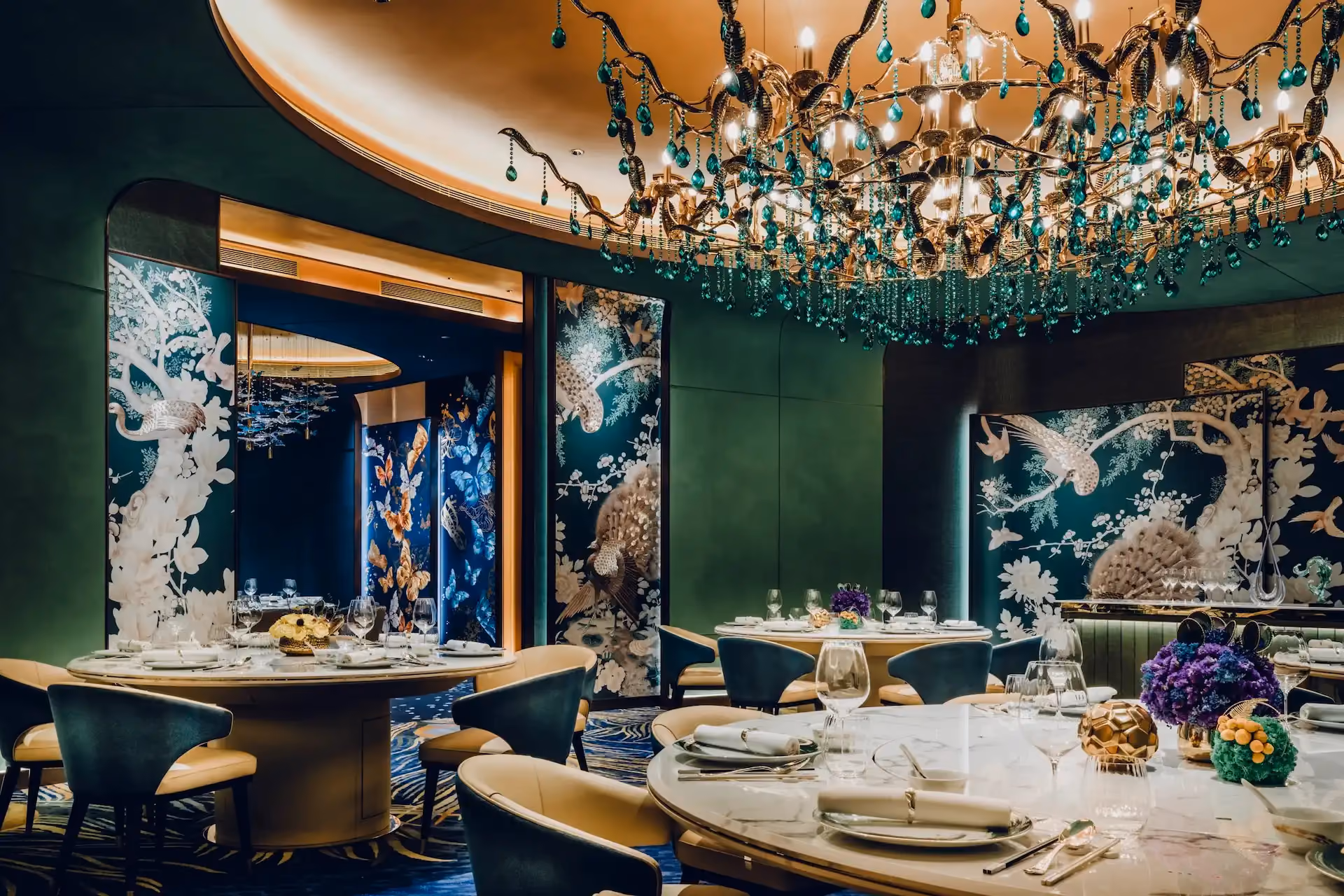 Grand Imperial Court private dining room Tapestry Of Feathers at MGM Macao featuring emerald crystal chandelier illuminated ceiling recess round marble banquet tables teal and cream upholstered chairs bespoke bird and floral wall murals and peacock feather patterned carpet within a luxury Chinese restaurant interior design