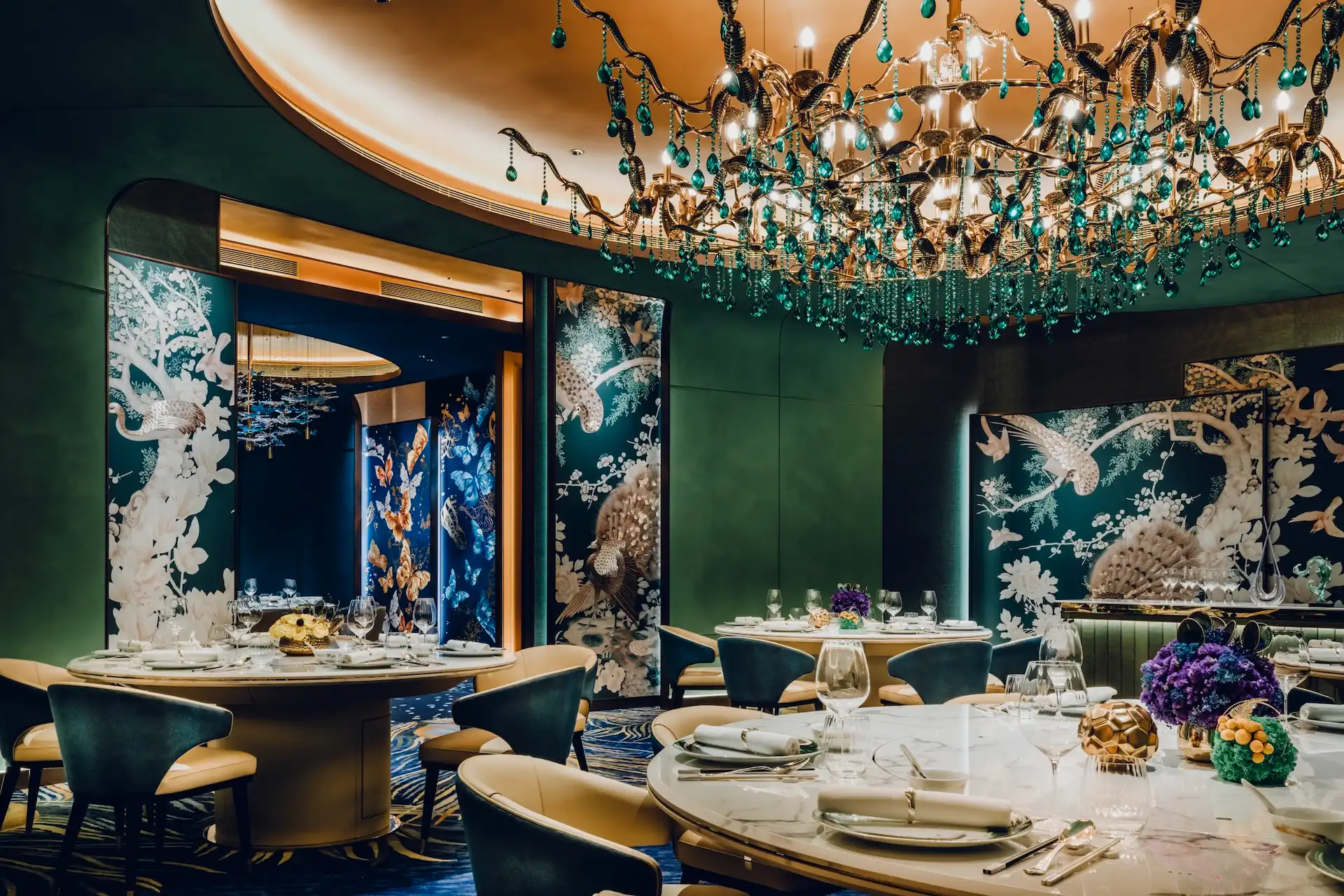 Grand Imperial Court private dining room Tapestry Of Feathers at MGM Macao featuring emerald crystal chandelier illuminated ceiling recess round marble banquet tables teal and cream upholstered chairs bespoke bird and floral wall murals and peacock feather patterned carpet within a luxury Chinese restaurant interior design