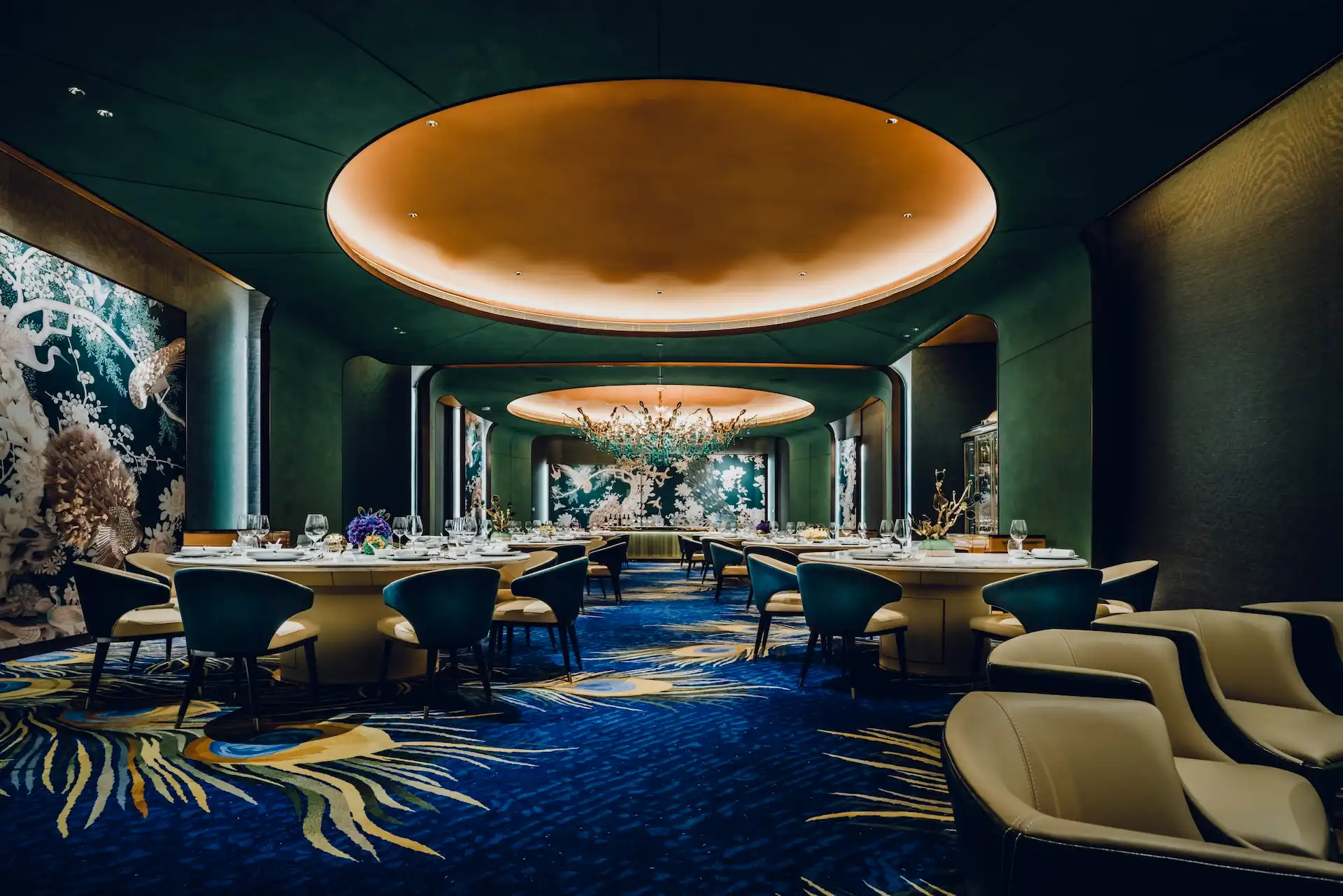 Grand Imperial Court Tapestry Of Feathers banquet dining interior at MGM Macao featuring symmetrical round dining tables teal upholstered chairs illuminated circular ceiling recess sculptural chandelier large scale bird and floral wall murals and bespoke peacock feather patterned carpet within a luxury Chinese restaurant design