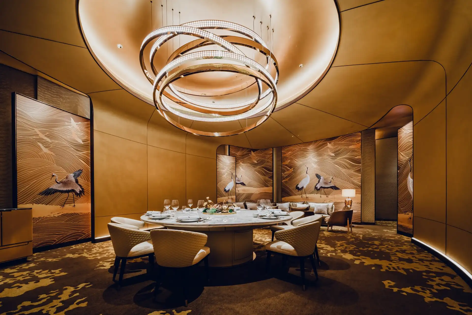 Grand Imperial Court private dining room at MGM Macao featuring sculptural circular chandelier suspended within recessed ceiling, round marble dining table with upholstered chairs and gold crane mural panels, defining a refined luxury Chinese restaurant interior design experience