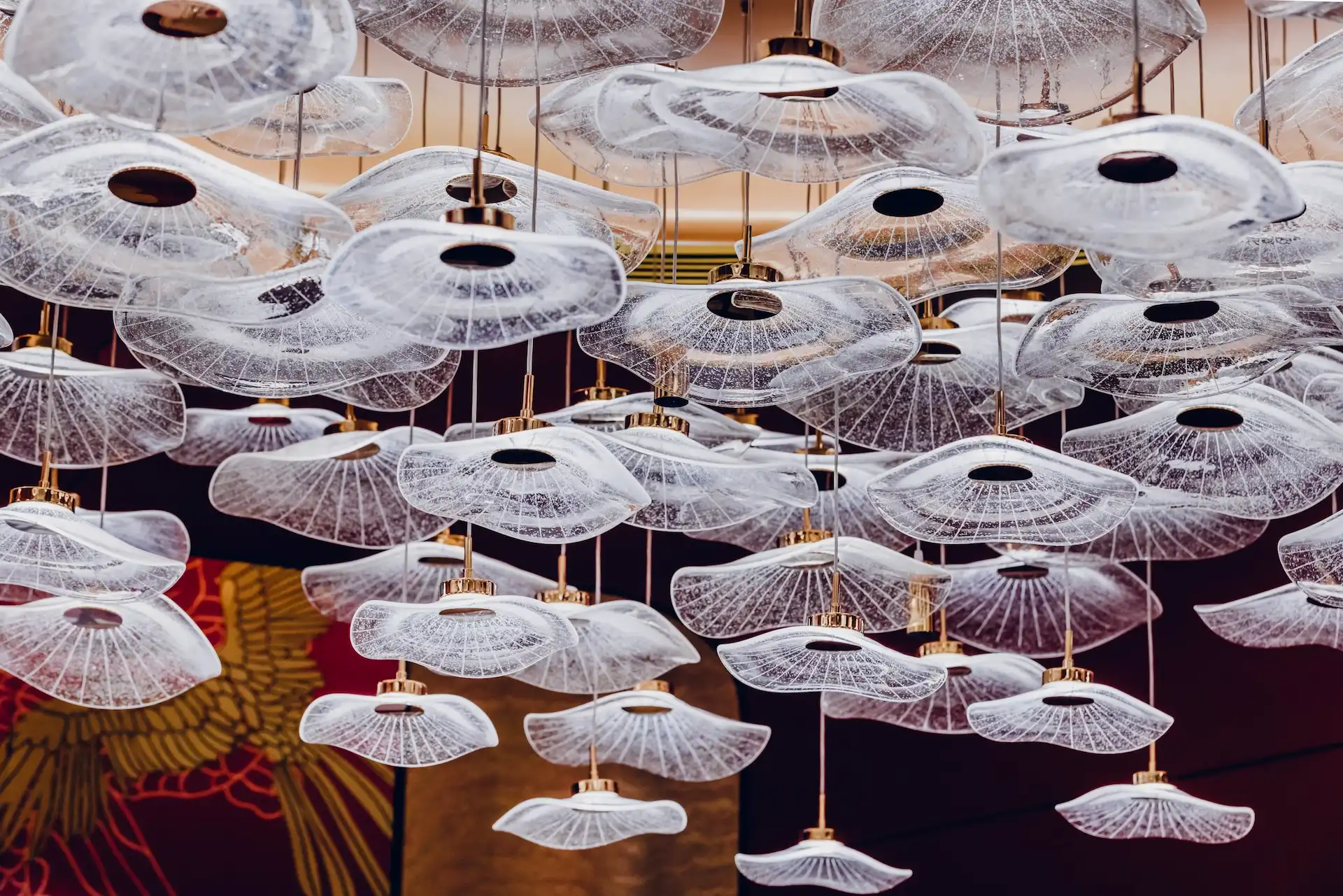 Grand Imperial Court Tapestry Of Parchment glass pendant detail at MGM Macao showing layered translucent glass discs with radial textures and brass fittings suspended at varying heights against red lacquer mural backdrop in a luxury Chinese restaurant interior