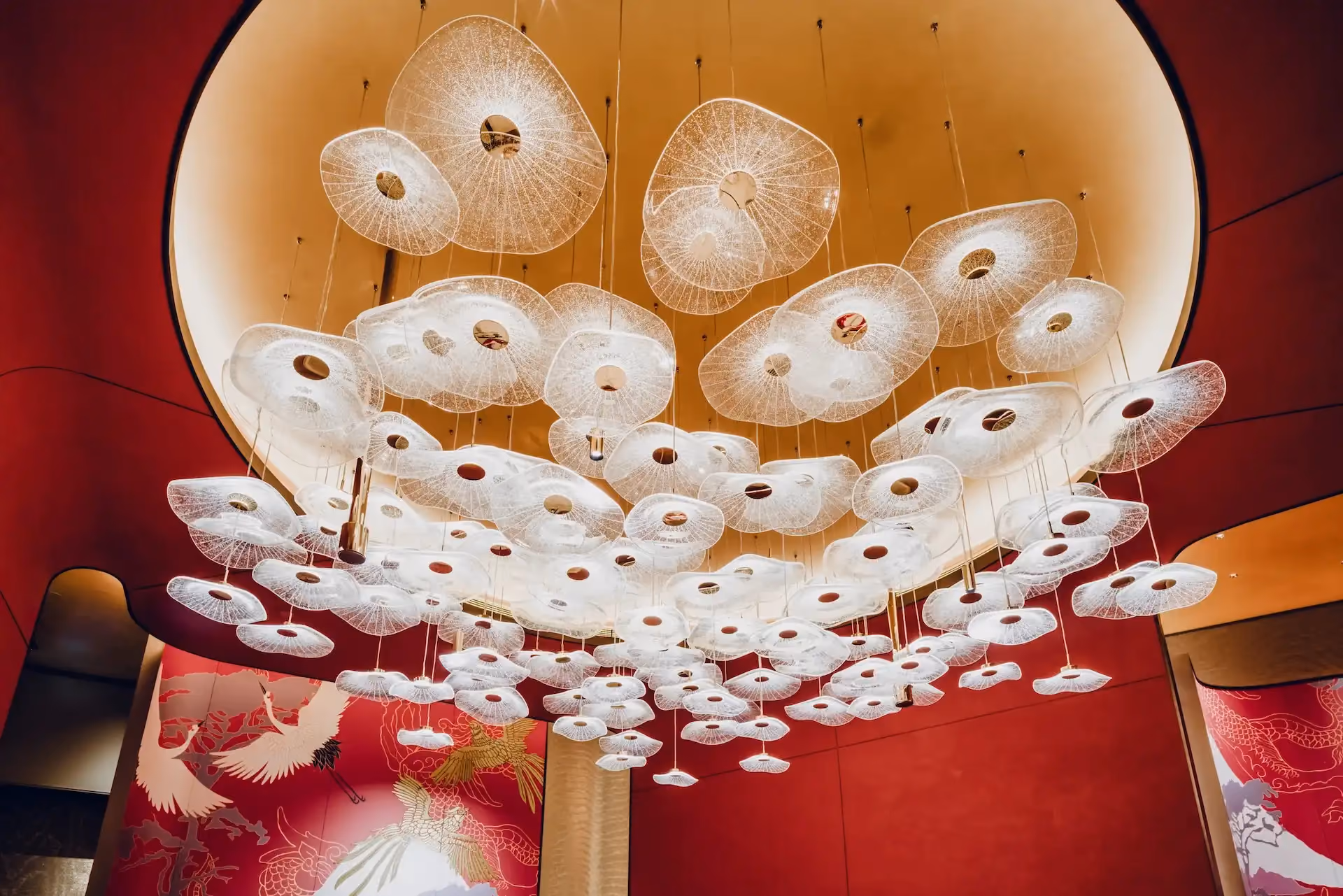 Grand Imperial Court Tapestry Of Parchment pendant light installation at MGM Macao featuring layered white disc pendants suspended within illuminated circular ceiling recess against red lacquer wall murals with gold phoenix motifs in a luxury Chinese restaurant interior design