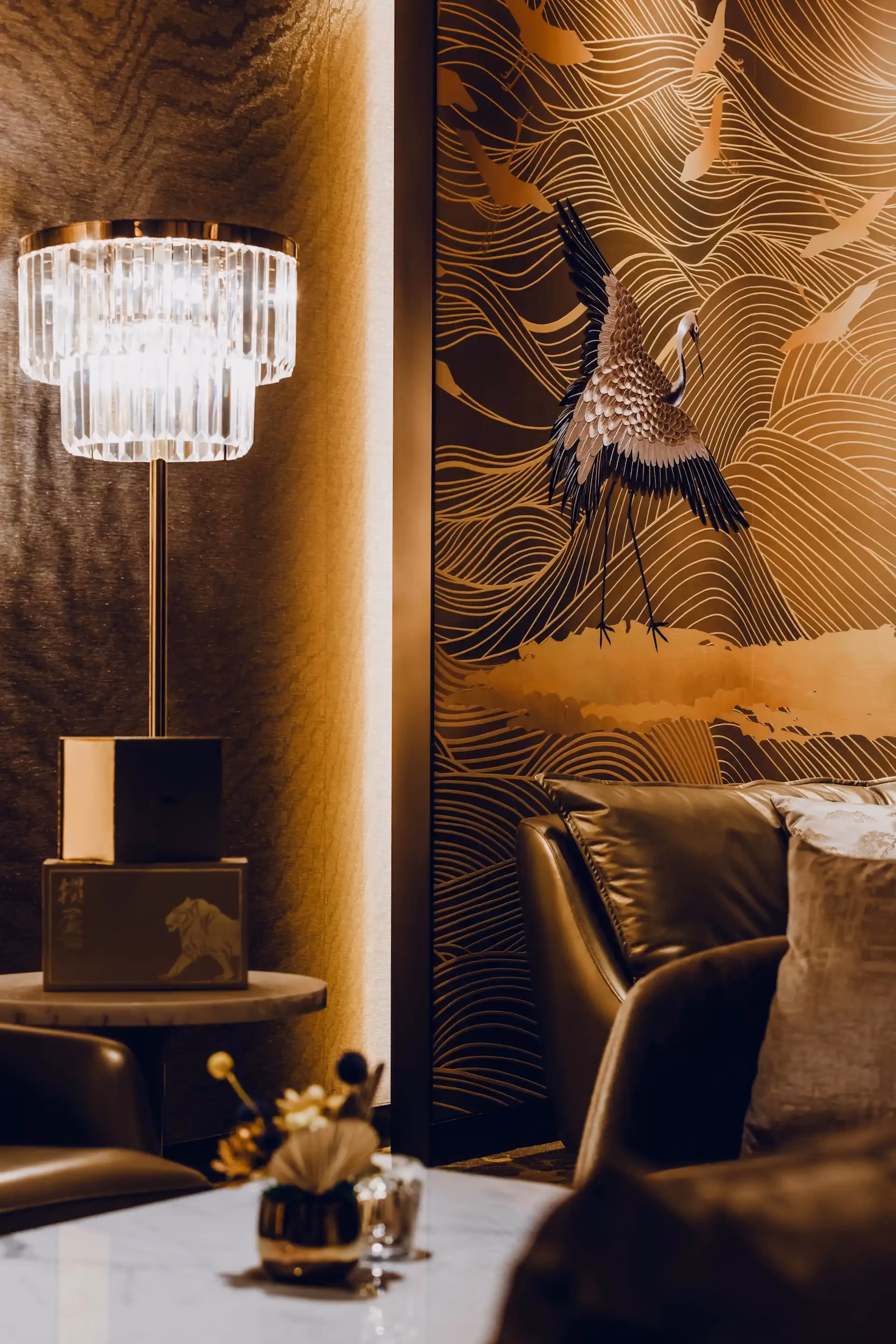 Detail of crystal floor lamp and leather lounge seating at Grand Imperial Court MGM Macau set against an illuminated crane wall panel with gold wave patterns and bronze textured wall finish.