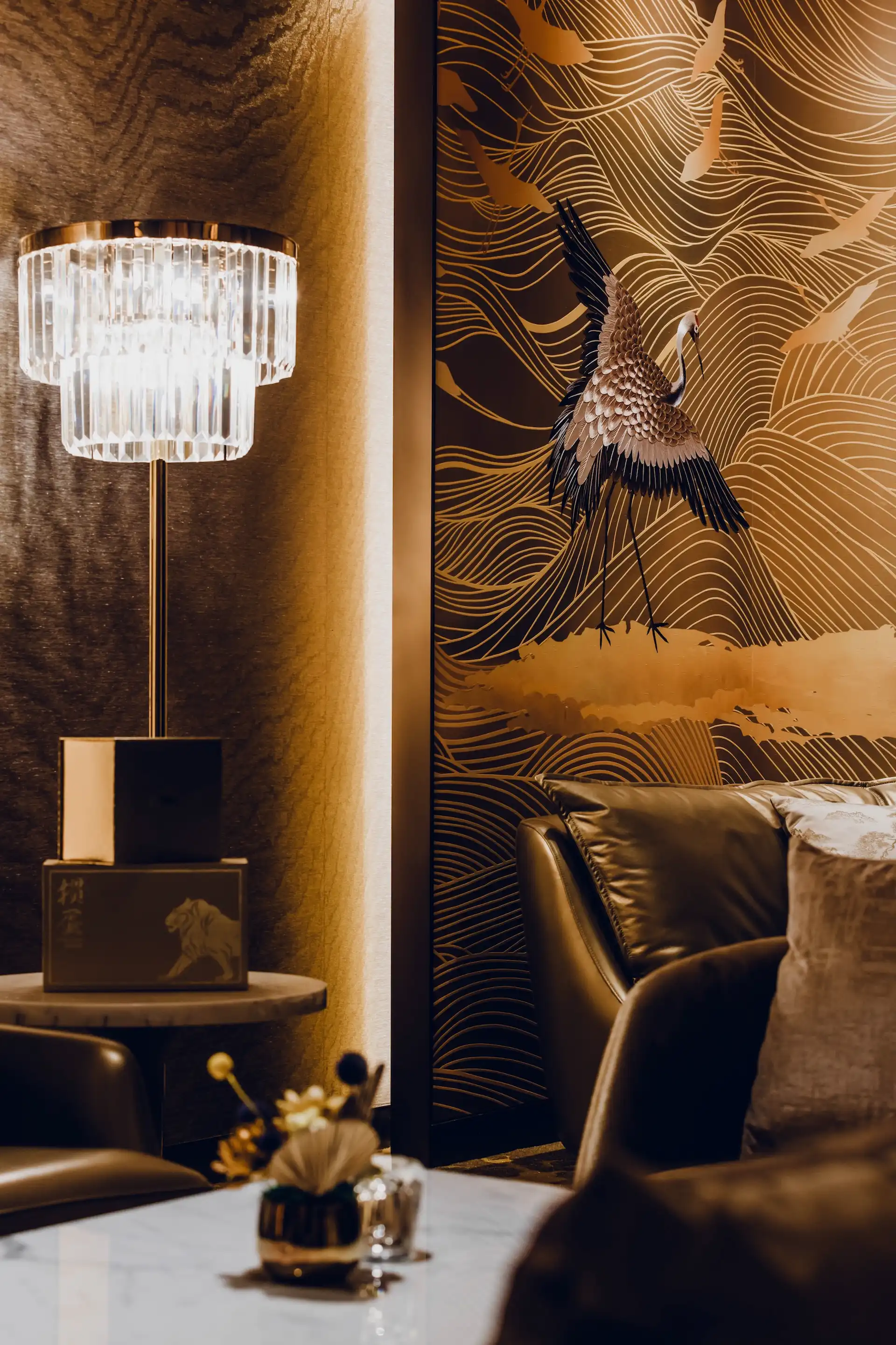 Detail of crystal floor lamp and leather lounge seating at Grand Imperial Court MGM Macau set against an illuminated crane wall panel with gold wave patterns and bronze textured wall finish.