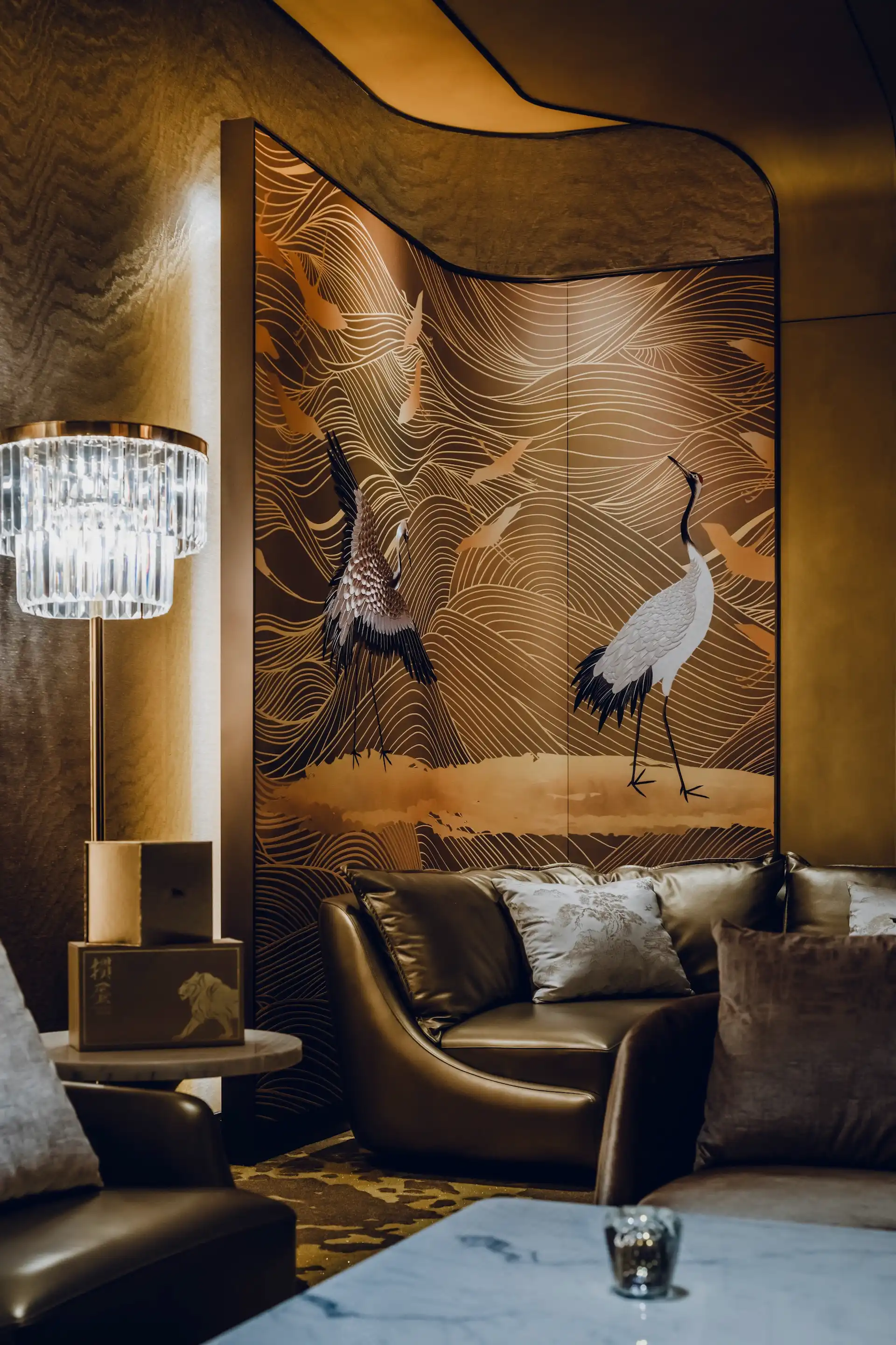 Lounge seating area at Grand Imperial Court MGM Macau with curved leather sofa, marble table and illuminated crane wall panel featuring gold wave patterns and bronze textured walls.