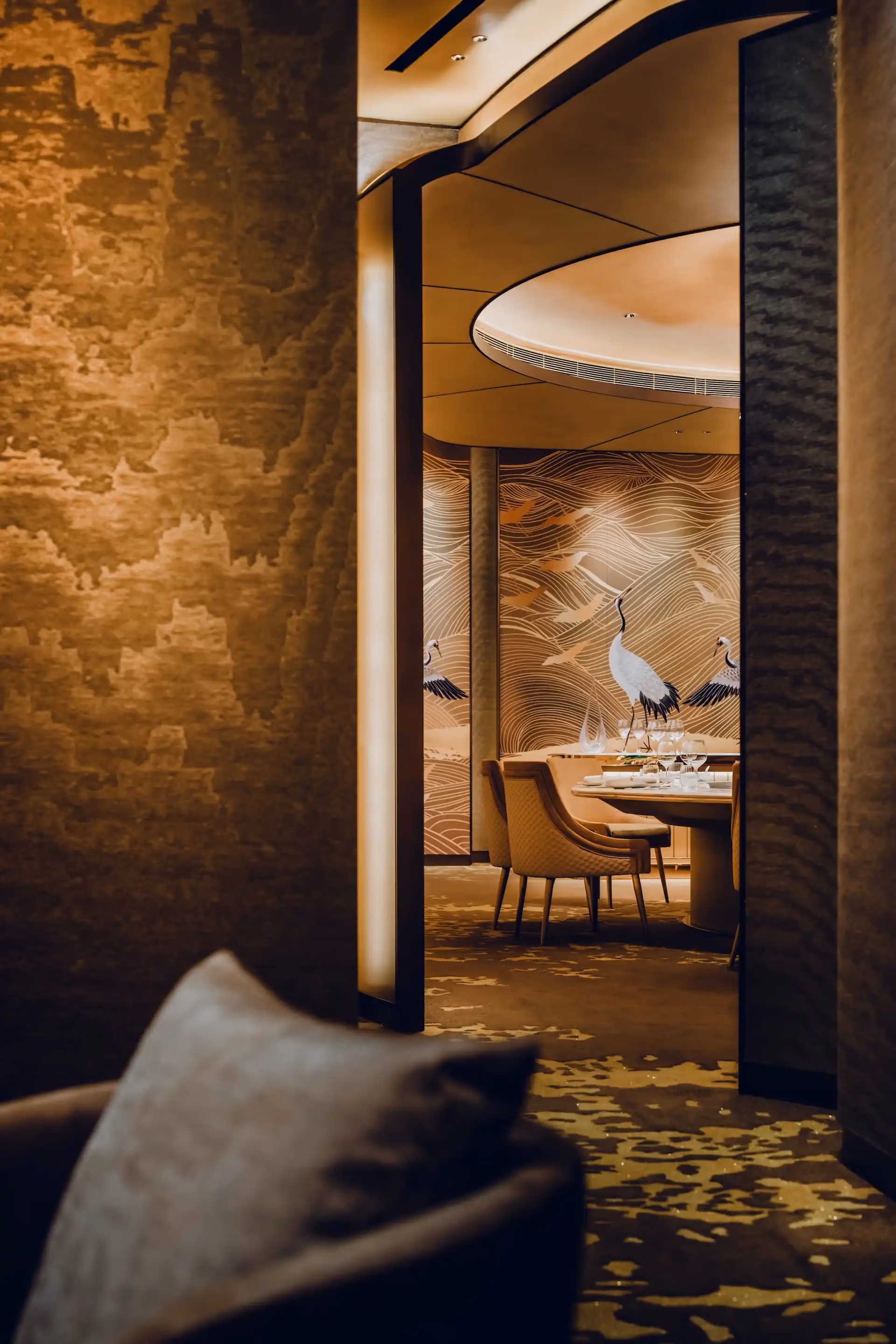 View through a bronze framed doorway at Grand Imperial Court MGM Macau leading to a private dining room with round table seating beneath a curved ceiling and crane wall panels with gold wave detailing.