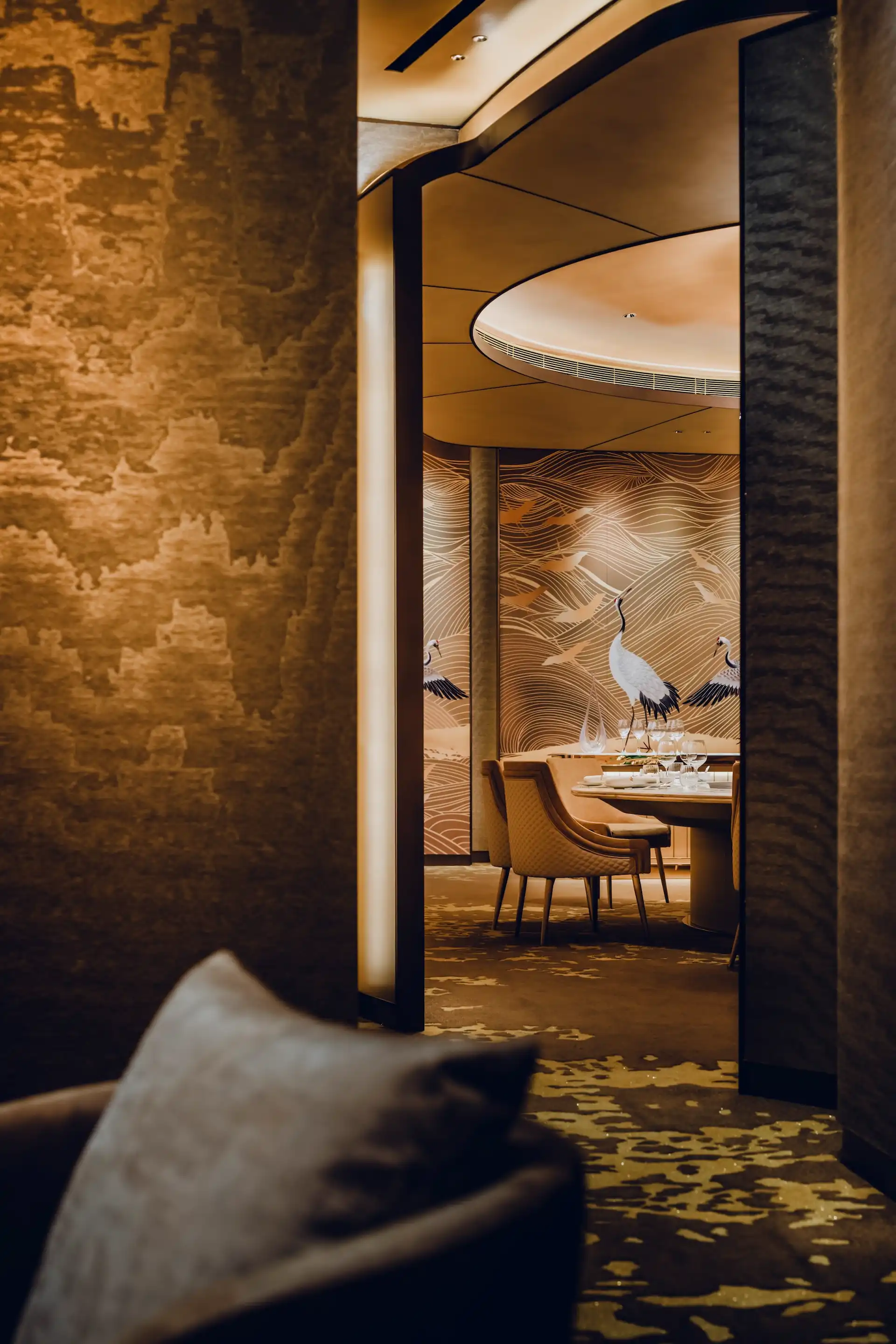View through a bronze framed doorway at Grand Imperial Court MGM Macau leading to a private dining room with round table seating beneath a curved ceiling and crane wall panels with gold wave detailing.