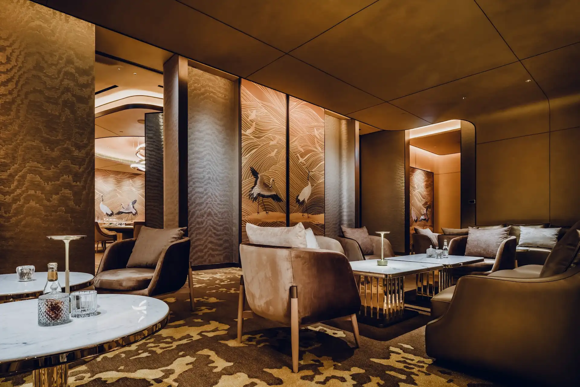 Lounge seating area at Grand Imperial Court MGM Macau with marble coffee tables, bronze metal bases, leather armchairs and backlit crane wall panels framed by textured bronze columns and curved ceiling details.