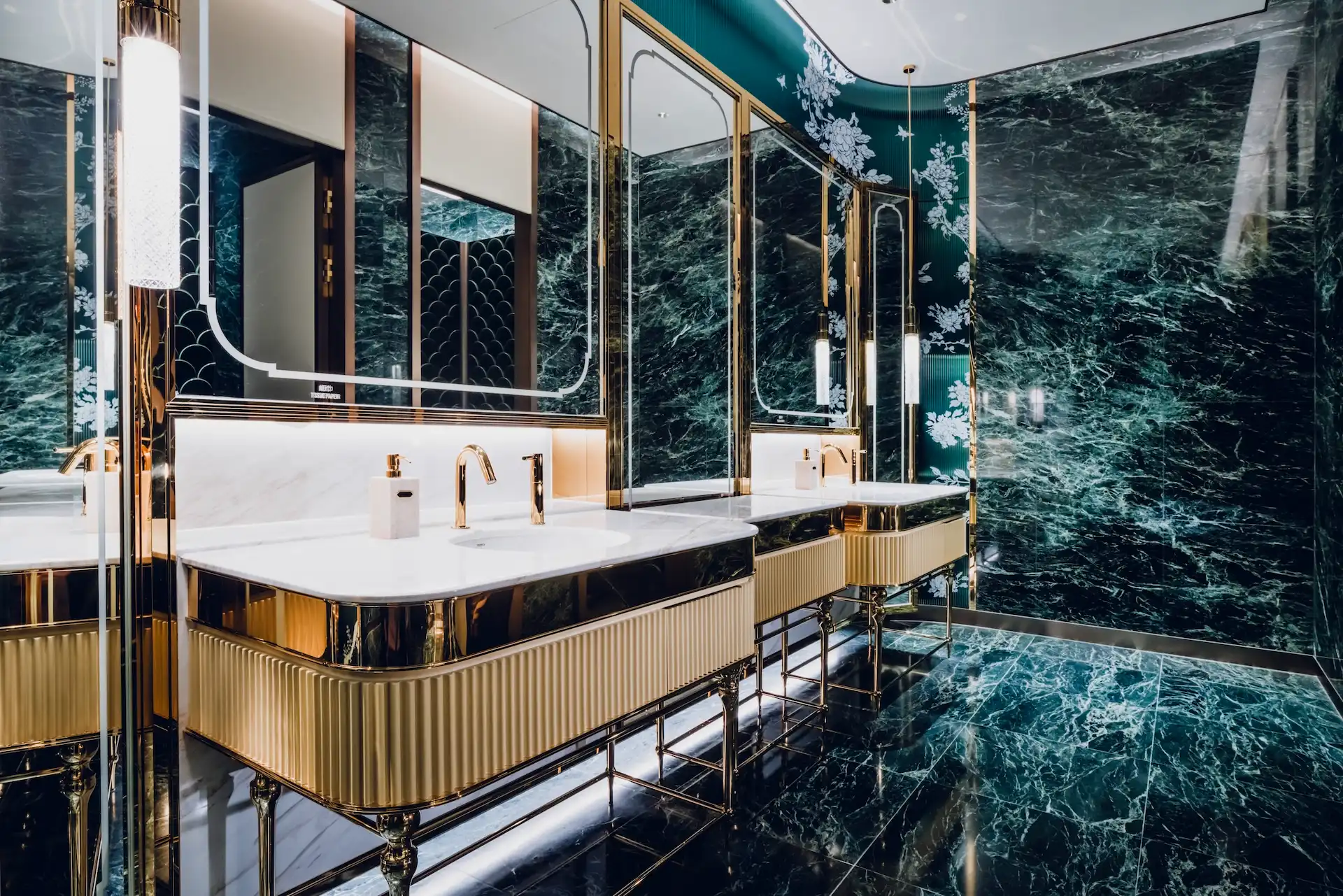 Restroom interior at Grand Imperial Court MGM Macau featuring dark green marble walls and floor, brass framed mirrors, fluted gold vanity units and green scalloped ceramic wall tiles as a feature backdrop.