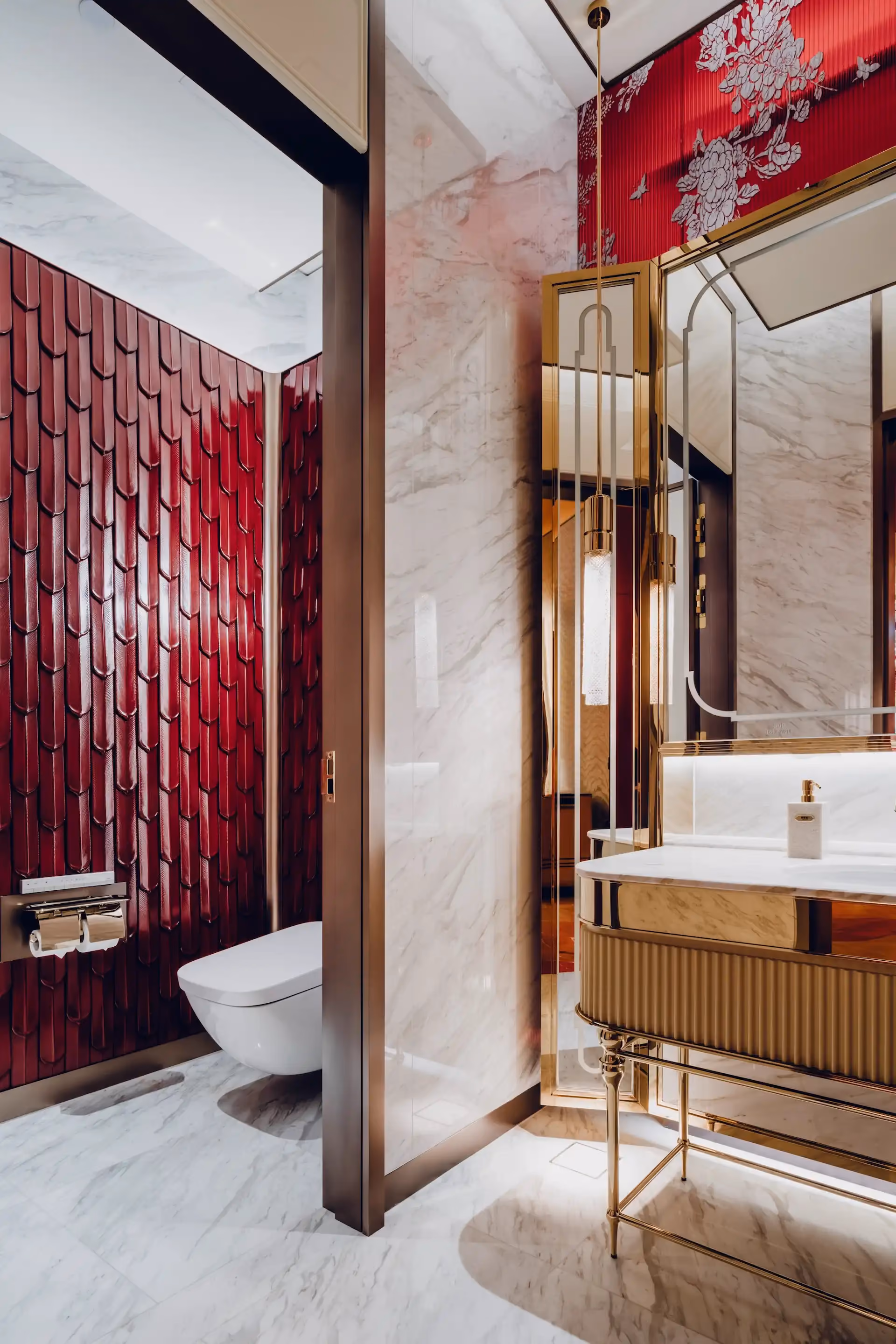 Restroom interior at Grand Imperial Court MGM Macau with white marble walls and floor, brass framed mirrors, fluted gold vanity unit and deep red sculpted ceramic wall tiles behind wall mounted toilet.