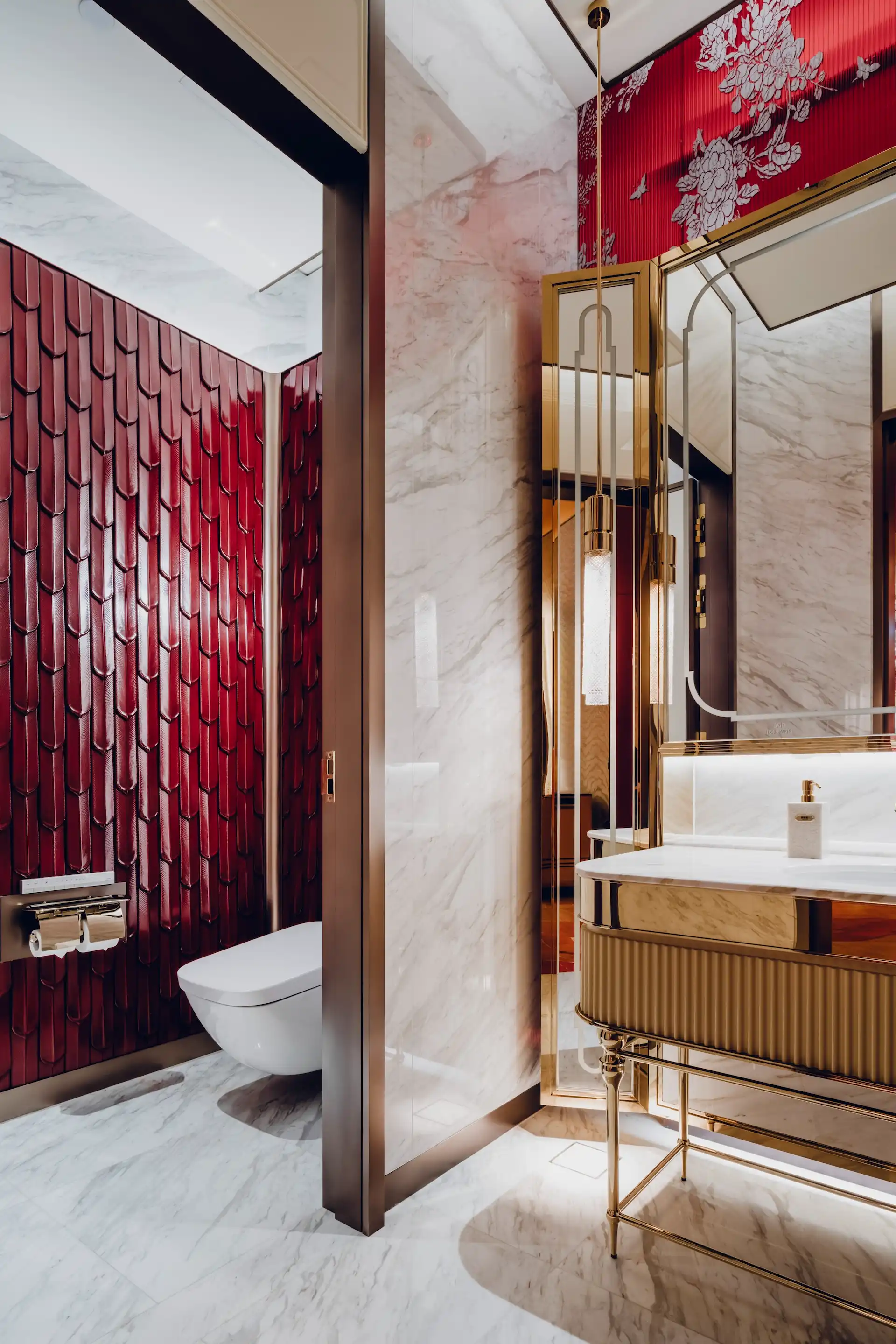 Restroom interior at Grand Imperial Court MGM Macau with white marble walls and floor, brass framed mirrors, fluted gold vanity unit and deep red sculpted ceramic wall tiles behind wall mounted toilet.