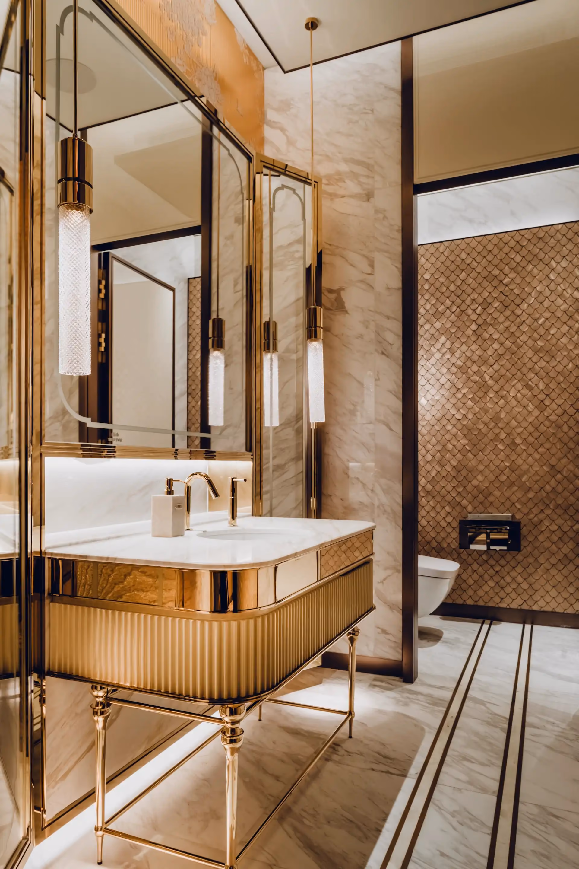 Restroom interior at Grand Imperial Court MGM Macau with marble vanity, polished gold fixtures, pendant lighting and metallic gold scalloped wall tiles behind wall mounted toilet.