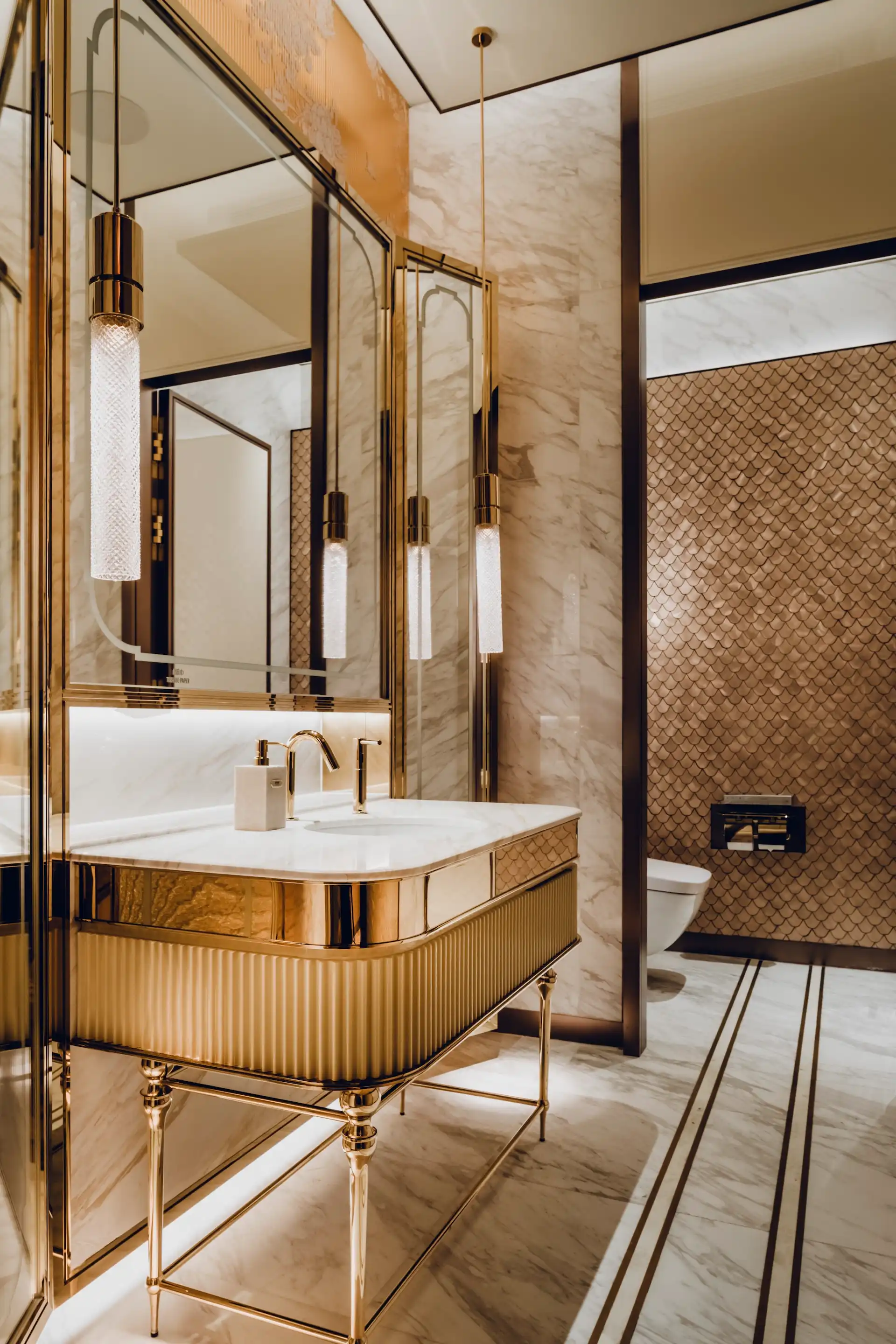 Restroom interior at Grand Imperial Court MGM Macau with marble vanity, polished gold fixtures, pendant lighting and metallic gold scalloped wall tiles behind wall mounted toilet.