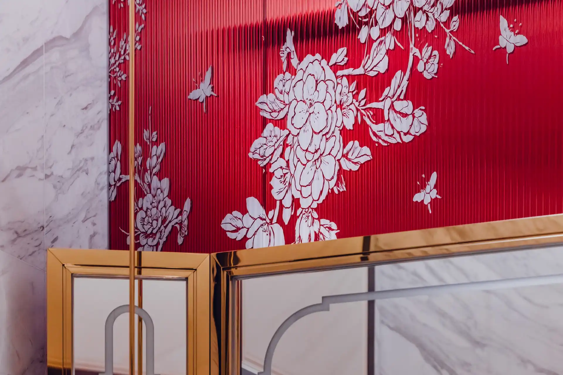 Mirror detail in Tapestry of Parchment restroom at Grand Imperial Court MGM Macau featuring brass framed mirror panels set against ribbed red glass wall with white floral motif and marble surrounds.