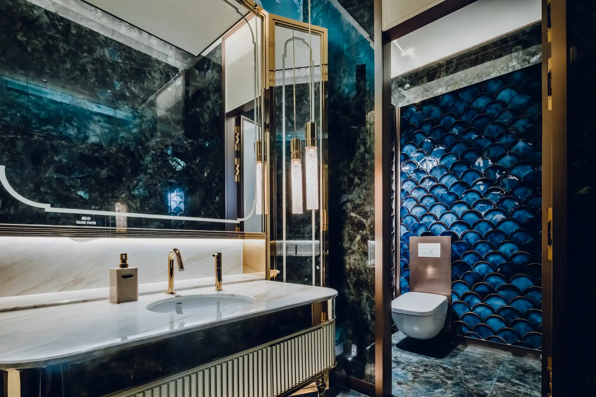 Restroom interior at Grand Imperial Court MGM Macau with marble vanity, gold fixtures, pendant lighting and deep blue scalloped wall tile behind wall mounted toilet.