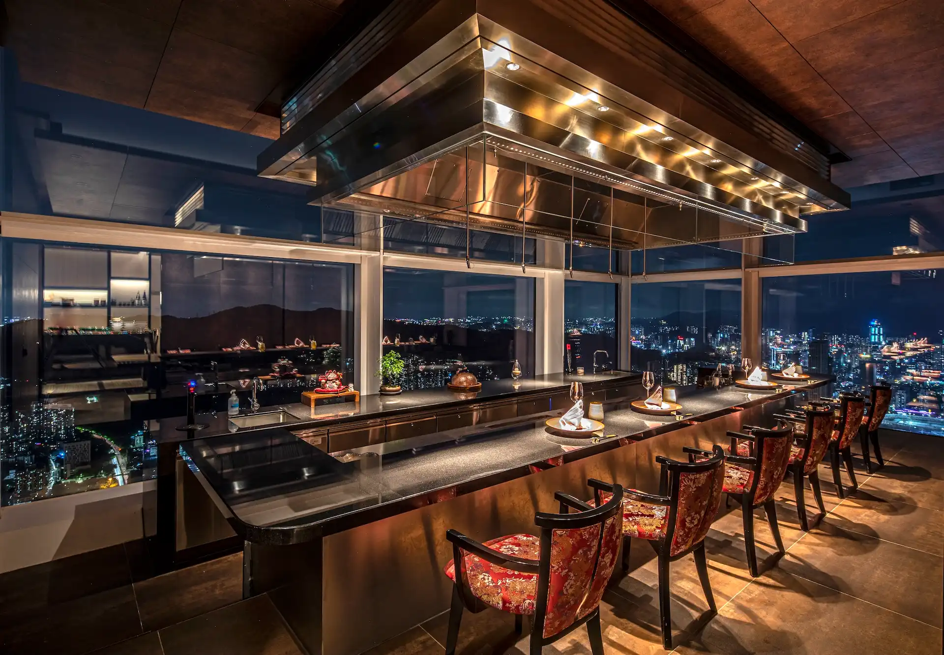 Private counter dining room at RIN Mandarin Oriental Shenzhen featuring teppanyaki style chef counter, corten steel finishes, suspended stainless steel hood, red upholstered chairs and panoramic nighttime city skyline views.