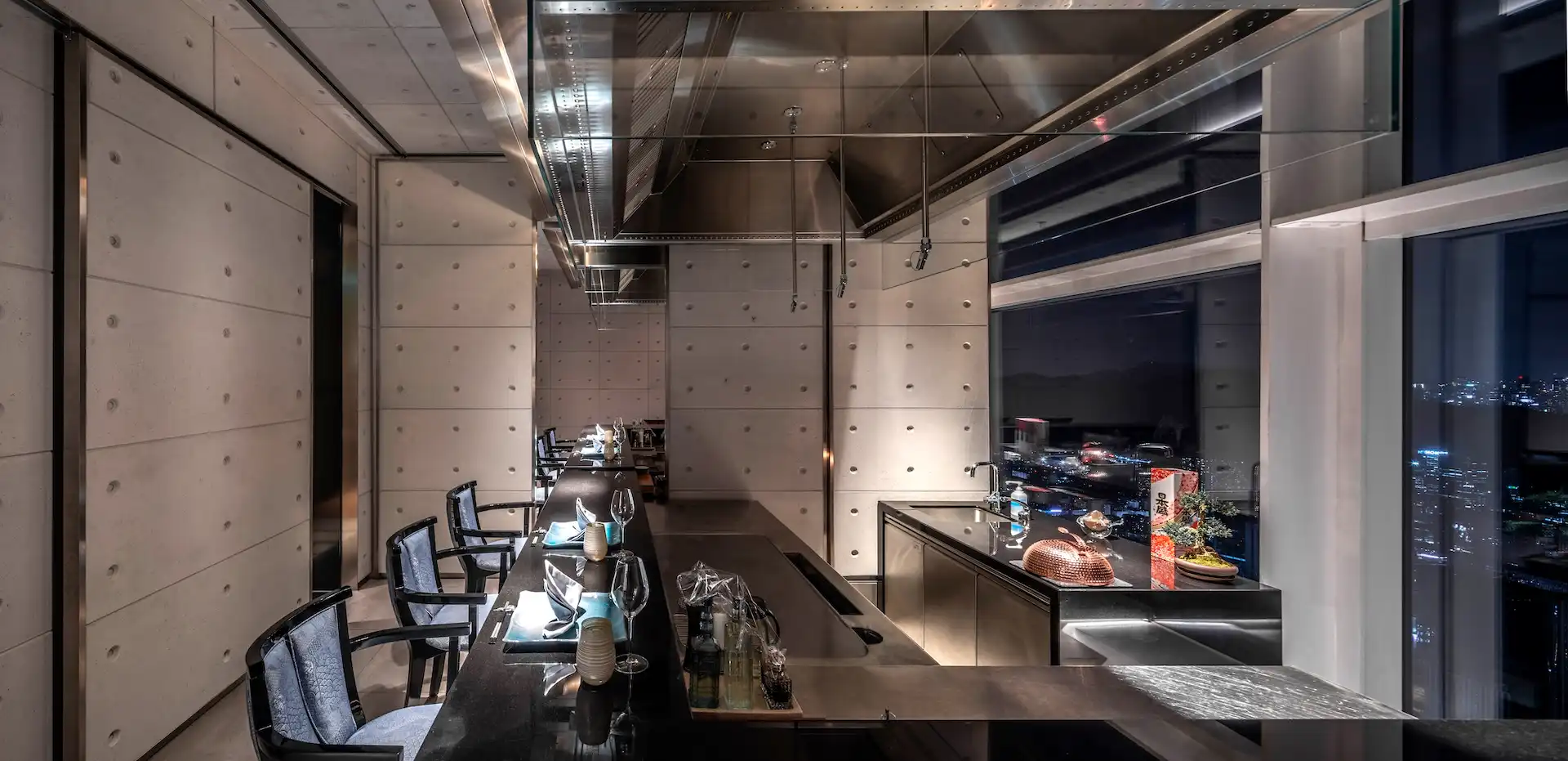 Concrete Room teppanyaki dining counter at RIN Mandarin Oriental Shenzhen featuring exposed concrete wall panels with recessed bolt detailing, a black granite chef station beneath a suspended stainless steel extraction hood, and floor to ceiling windows overlooking the Shenzhen skyline at night.