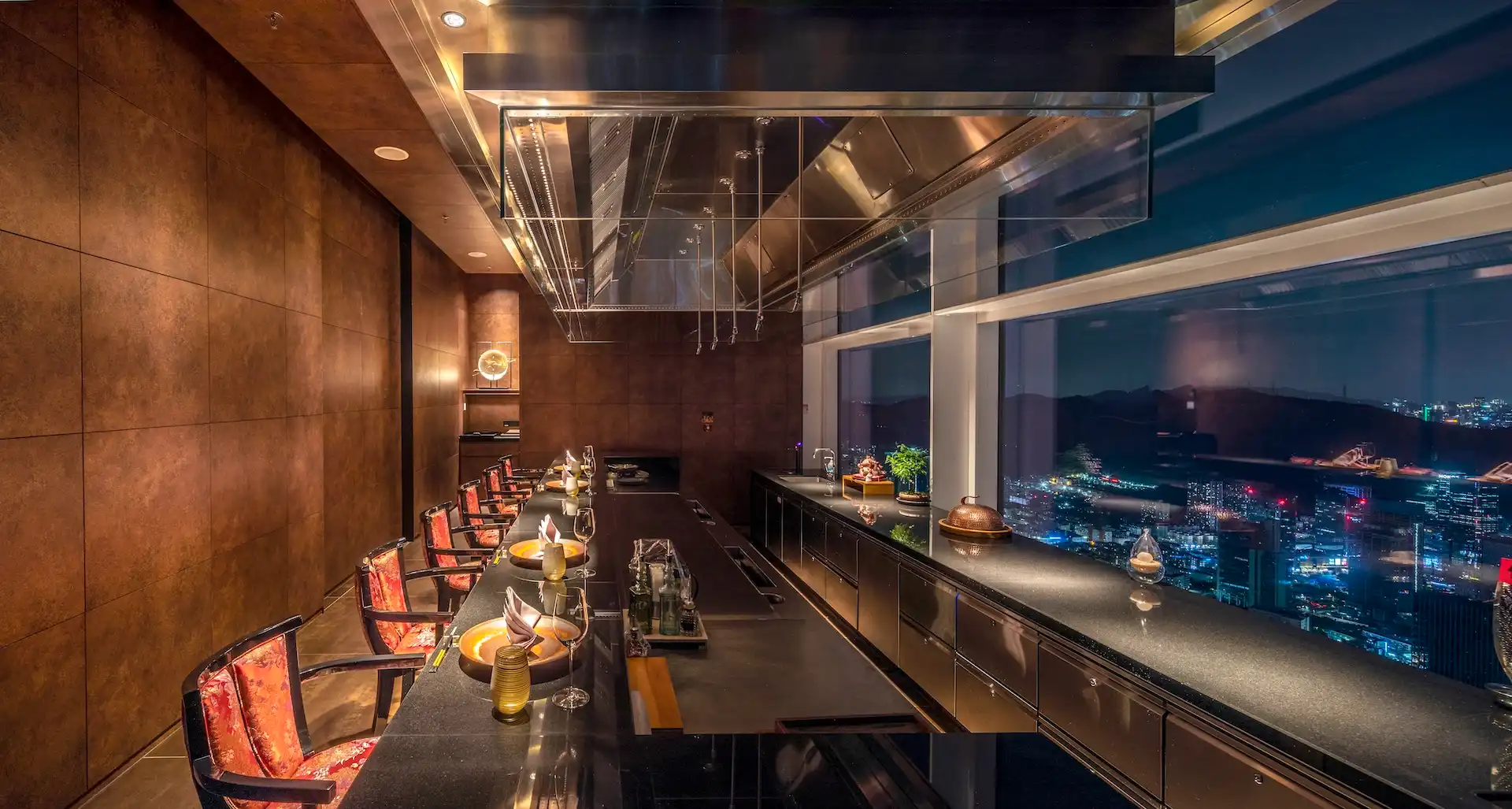 Teppanyaki private dining room at RIN Mandarin Oriental Shenzhen featuring long black stone chef counter, corten steel wall panels, suspended stainless steel extraction hood, red patterned dining chairs and panoramic night views of the Shenzhen skyline.
