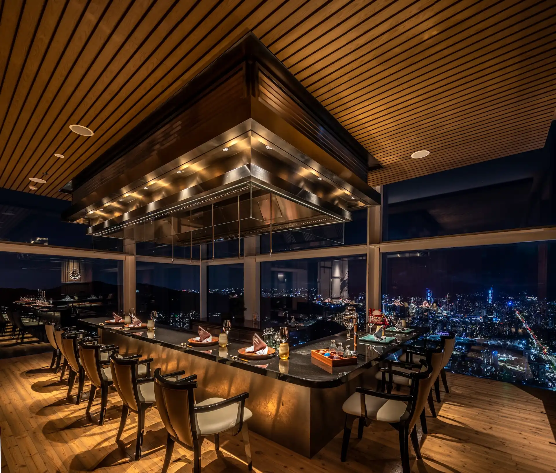 Pine Room teppanyaki dining counter at RIN Mandarin Oriental Shenzhen featuring a granite chef station beneath a suspended stainless steel extraction hood, warm pine timber ceiling and flooring, and panoramic floor to ceiling windows overlooking the Shenzhen night skyline.