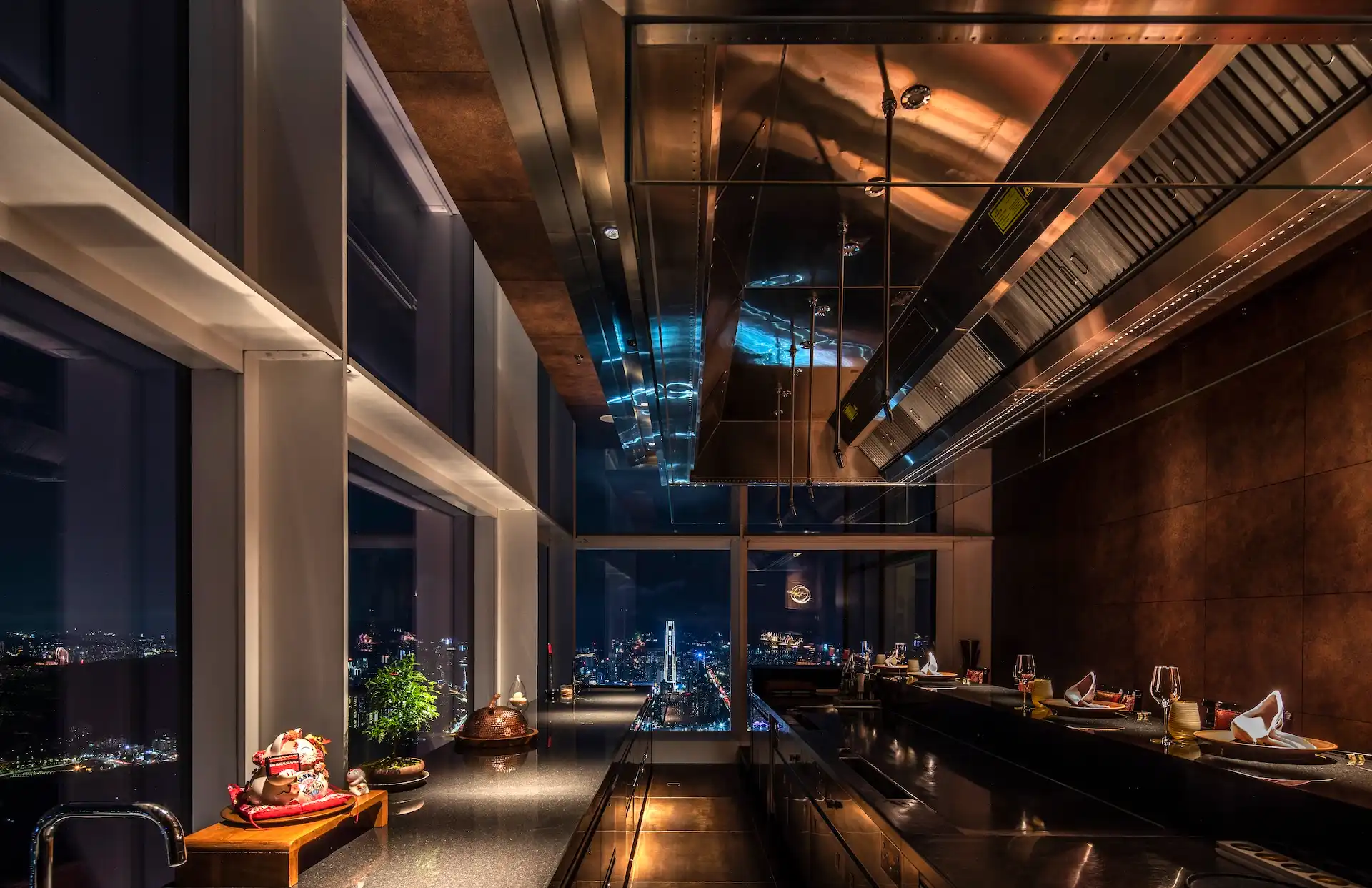 Teppanyaki counter detail at RIN Mandarin Oriental Shenzhen featuring polished steel cooking hood, black stone counter top, corten steel wall panels and panoramic city skyline views through floor to ceiling glazing at night.