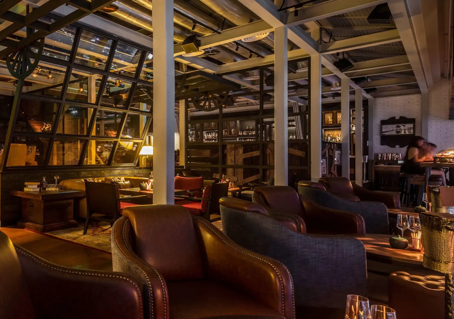 Wide interior view of Prohibition Bar Mandarin Oriental Shenzhen showing leather lounge armchairs, timber tables with candlelight, steel-framed glazing partitions, exposed structural columns and ductwork, layered shelving displays, and warm ambient lighting defining an intimate industrial speakeasy setting.