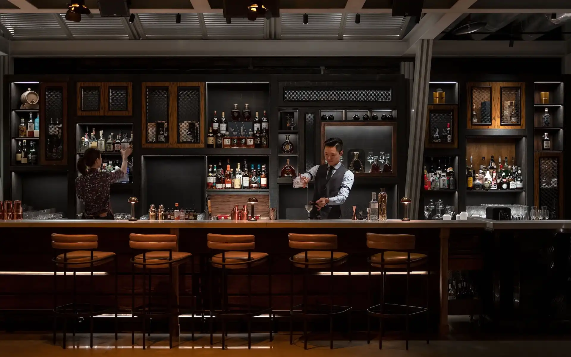 Main bar counter at Prohibition Bar Mandarin Oriental Shenzhen featuring bartenders preparing cocktails behind a timber bar, illuminated back bar shelving with premium spirits display, leather bar stools, and a warm speakeasy inspired atmosphere.