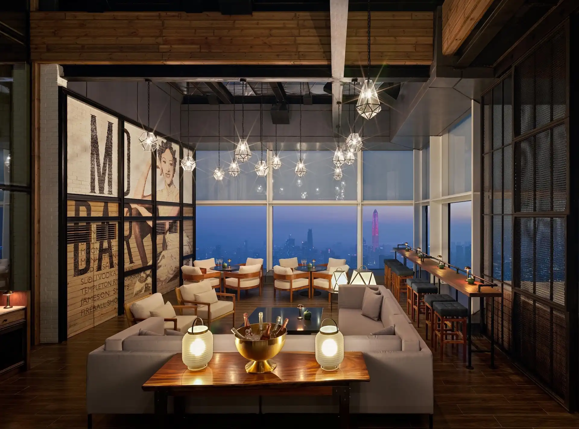 Window lounge at Prohibition Bar Mandarin Oriental Shenzhen featuring upholstered sofas, timber lounge chairs and low tables, pendant cluster lighting, a high counter with bar stools along the glazing, large-scale graphic wall art, and panoramic city skyline views at dusk.