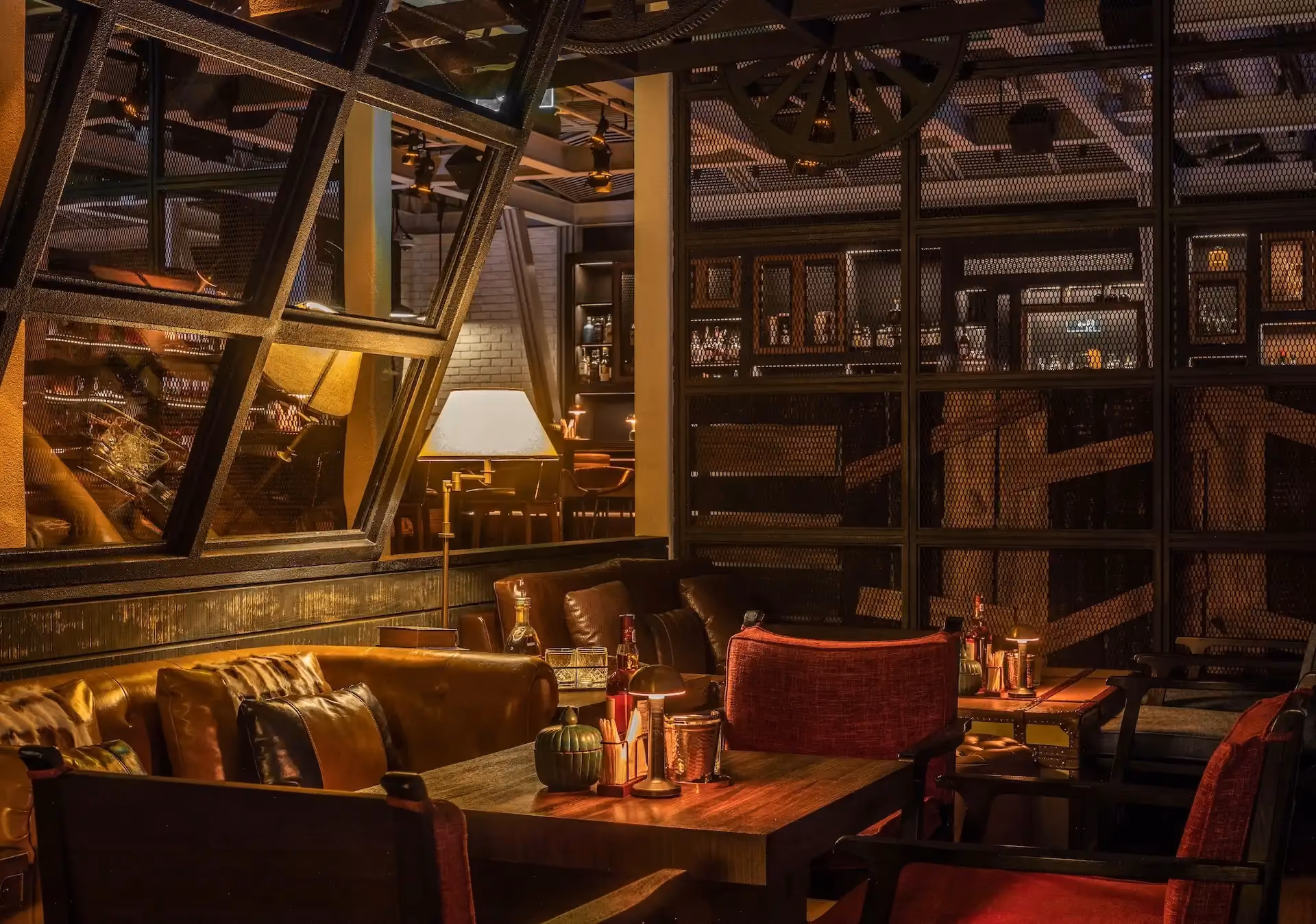 Detailed view of Prohibition Bar Mandarin Oriental Shenzhen lounge seating with leather sofas, red upholstered armchairs, timber cocktail tables with candlelight and whisky service, steel-framed mesh partitions, angled industrial glazing, and warm layered lighting creating an intimate speakeasy setting.
