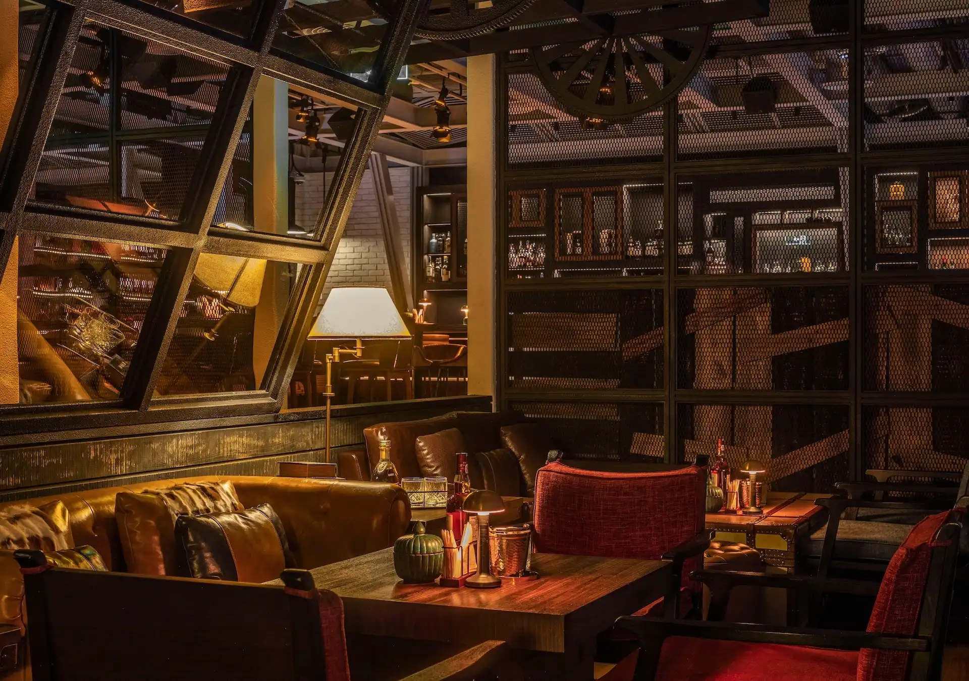 Detailed view of Prohibition Bar Mandarin Oriental Shenzhen lounge seating with leather sofas, red upholstered armchairs, timber cocktail tables with candlelight and whisky service, steel-framed mesh partitions, angled industrial glazing, and warm layered lighting creating an intimate speakeasy setting.