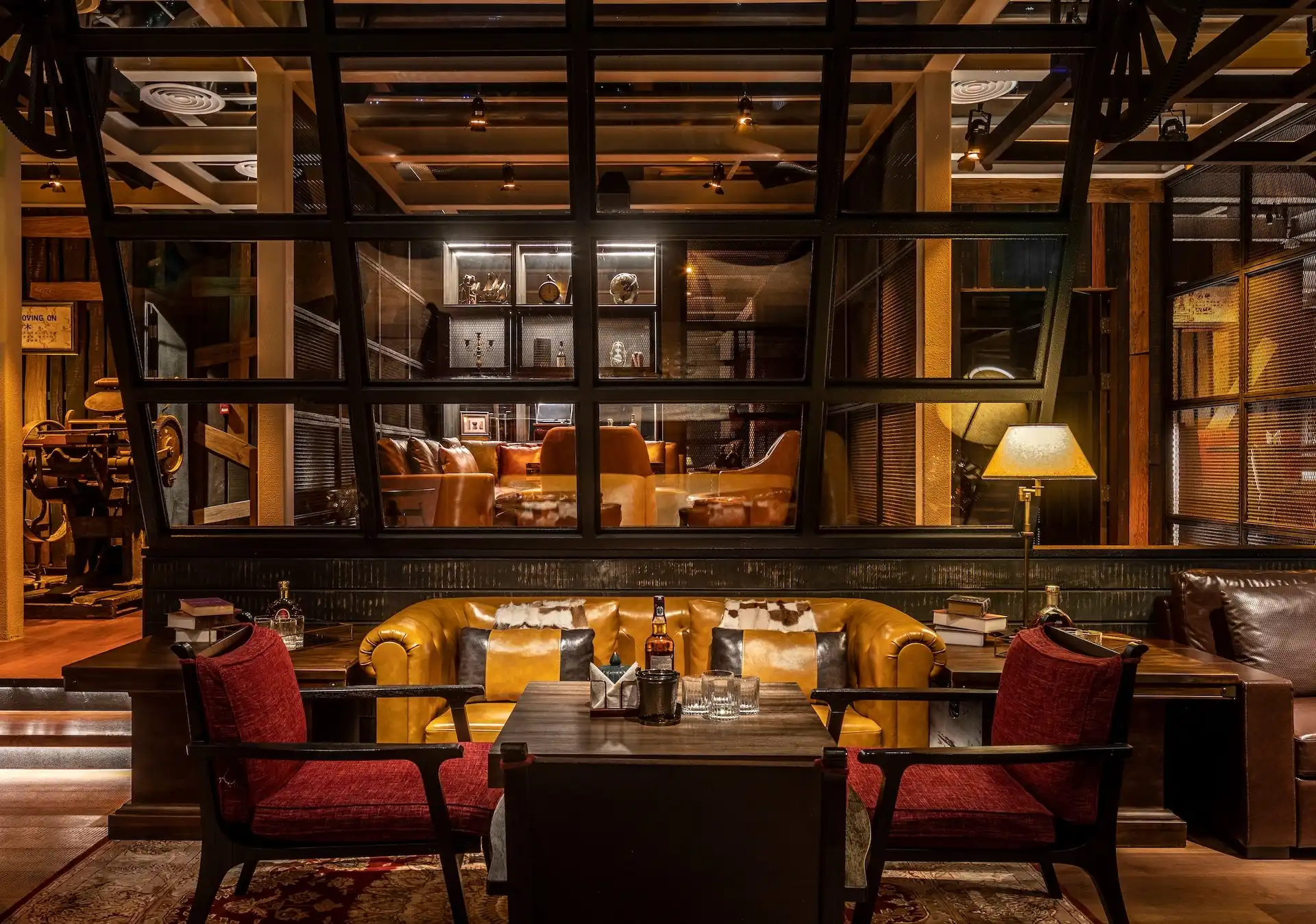 Lounge seating area at Prohibition Bar Mandarin Oriental Shenzhen featuring leather Chesterfield sofas, red upholstered armchairs, timber coffee tables with whisky service, layered shelving displays behind steel-framed glazing, warm table lamps, and a richly textured rug enhancing the intimate speakeasy atmosphere.